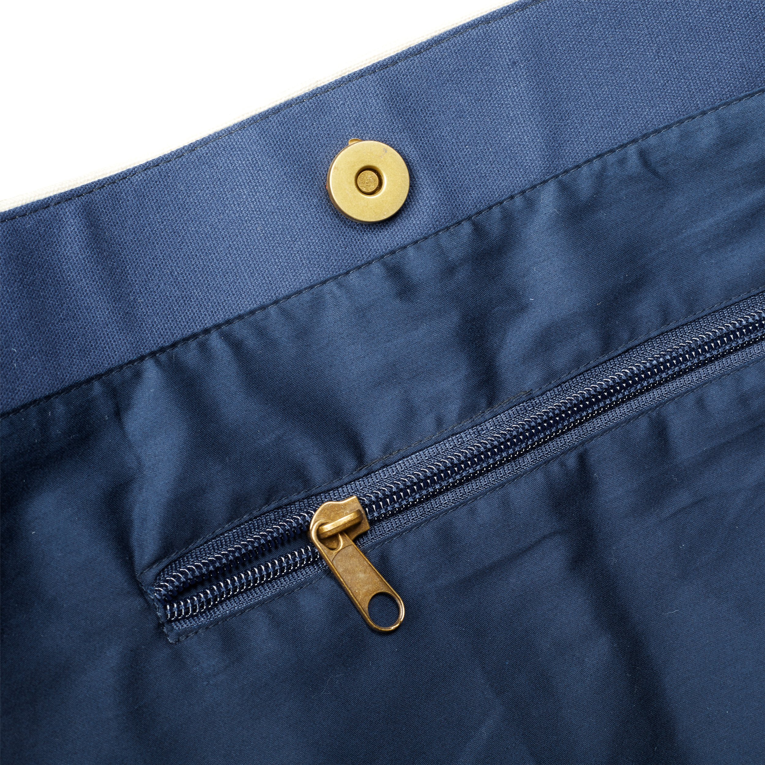 Close-up of the interior of a navy blue bag, showing a zippered pocket with a brass zipper and a magnetic snap button closure. The fabric has a smooth, slightly shiny texture.