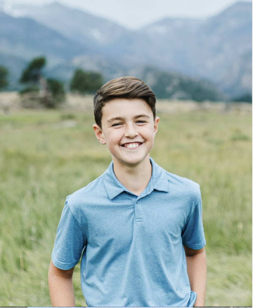 A smiling young boy with dark brown hair wearing a blue polo shirt standing in a green field with mountains in the background.