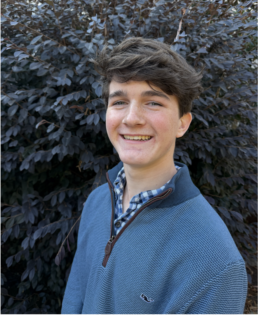 A young man with wavy brown hair and blue eyes smiling, standing outdoors in front of dark purple leaves.