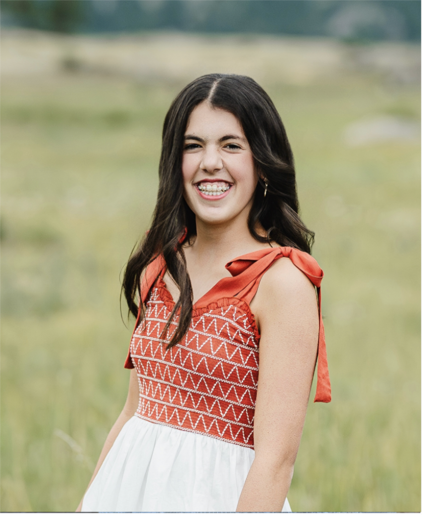 A young girl with long dark hair smiling and winking outdoors in a green field, wearing a red and white dress with shoulder bows.