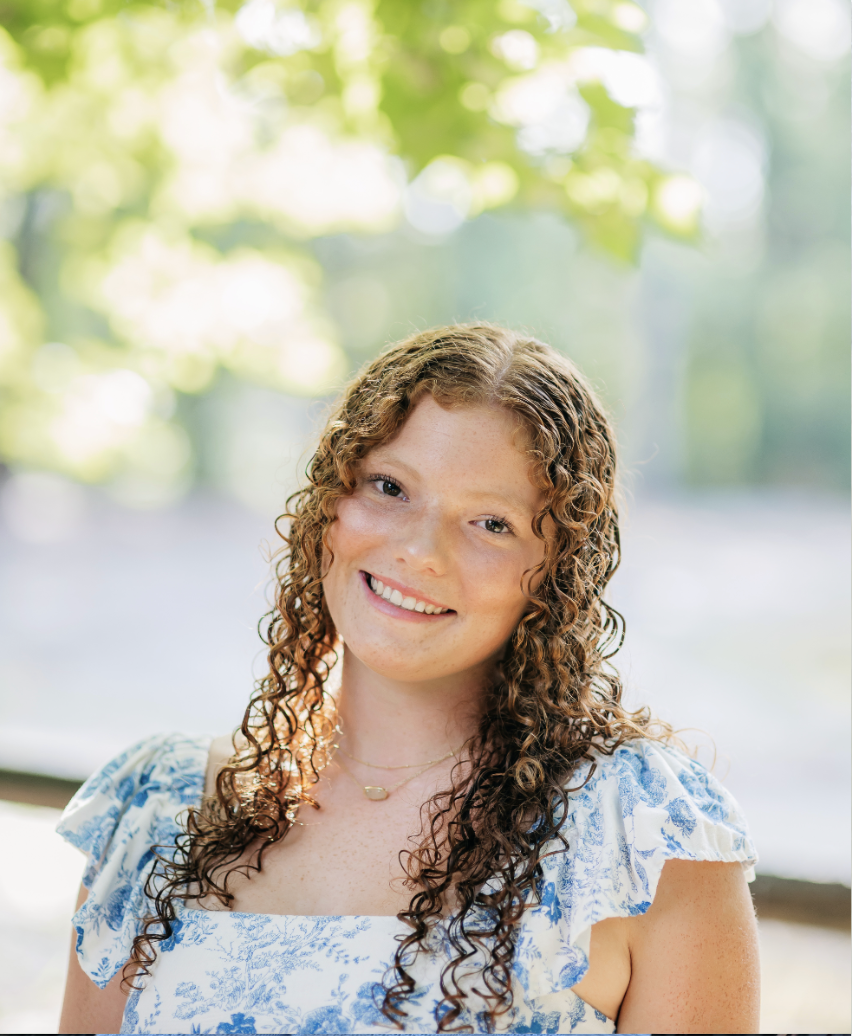 A young woman with curly red hair and freckles, smiling outdoors with green blurred trees in the background, wearing a blue and white floral dress.