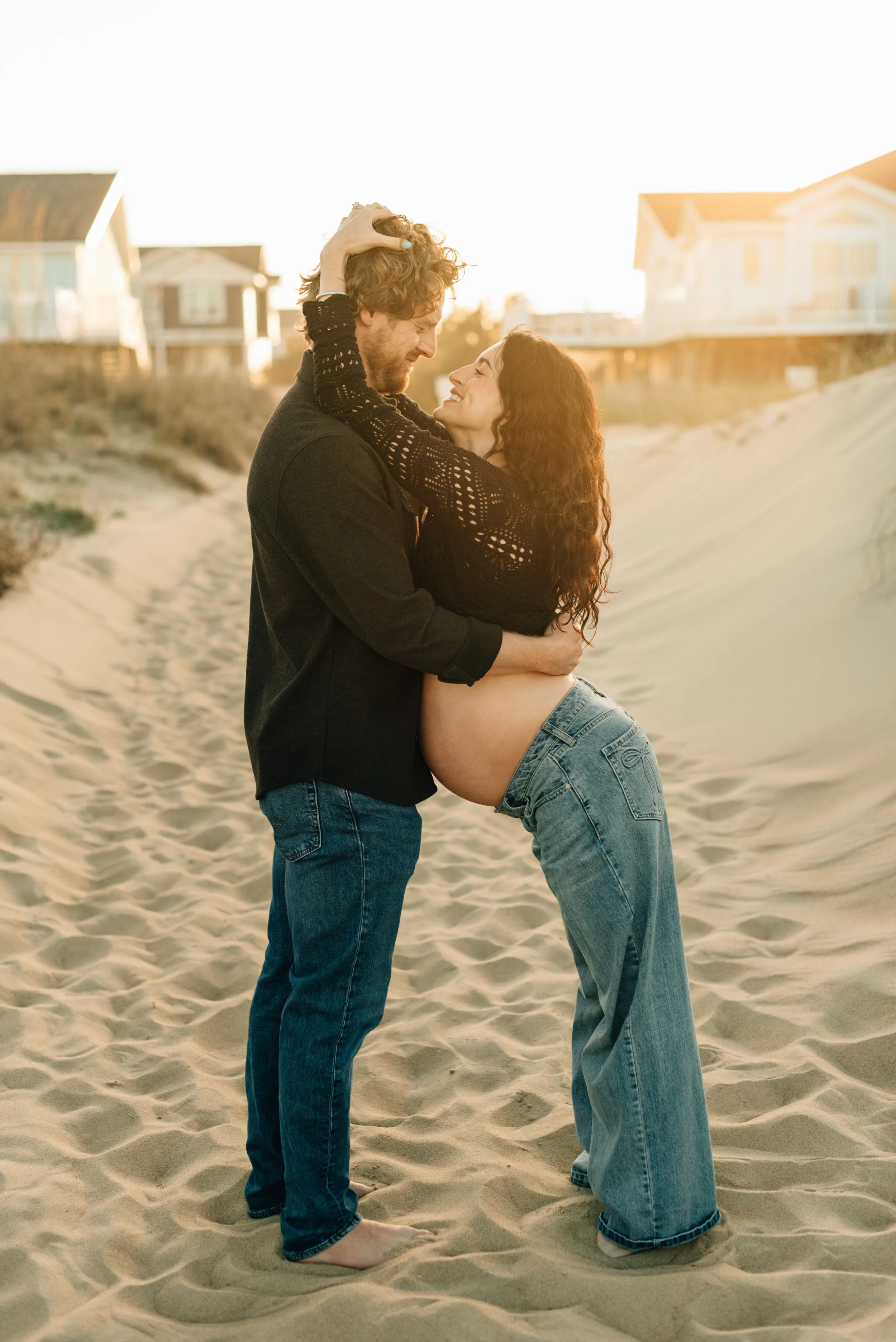 Pregnant woman and partner standing on a sandy beach with tall grass.