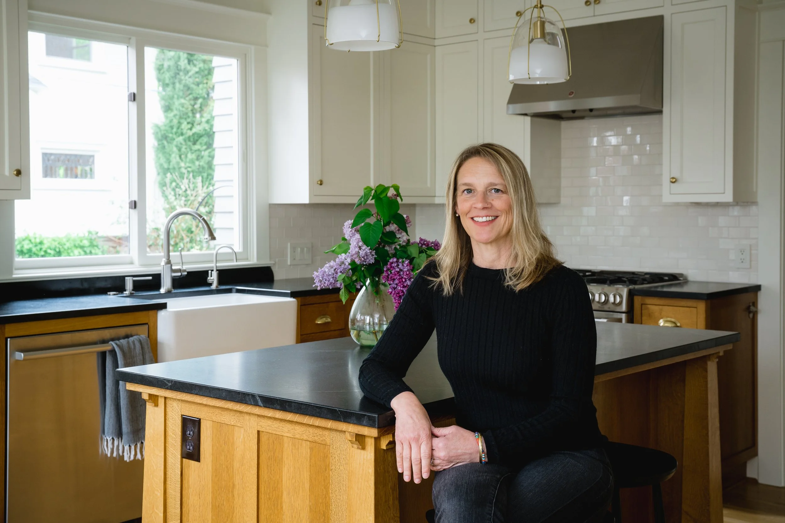 A professional woman in upscale kitchen with a friendly smile. The kitchen features stylish white cabinets, a window, a flower vase with purple flowers, a farmhouse sink, and a professional stove.