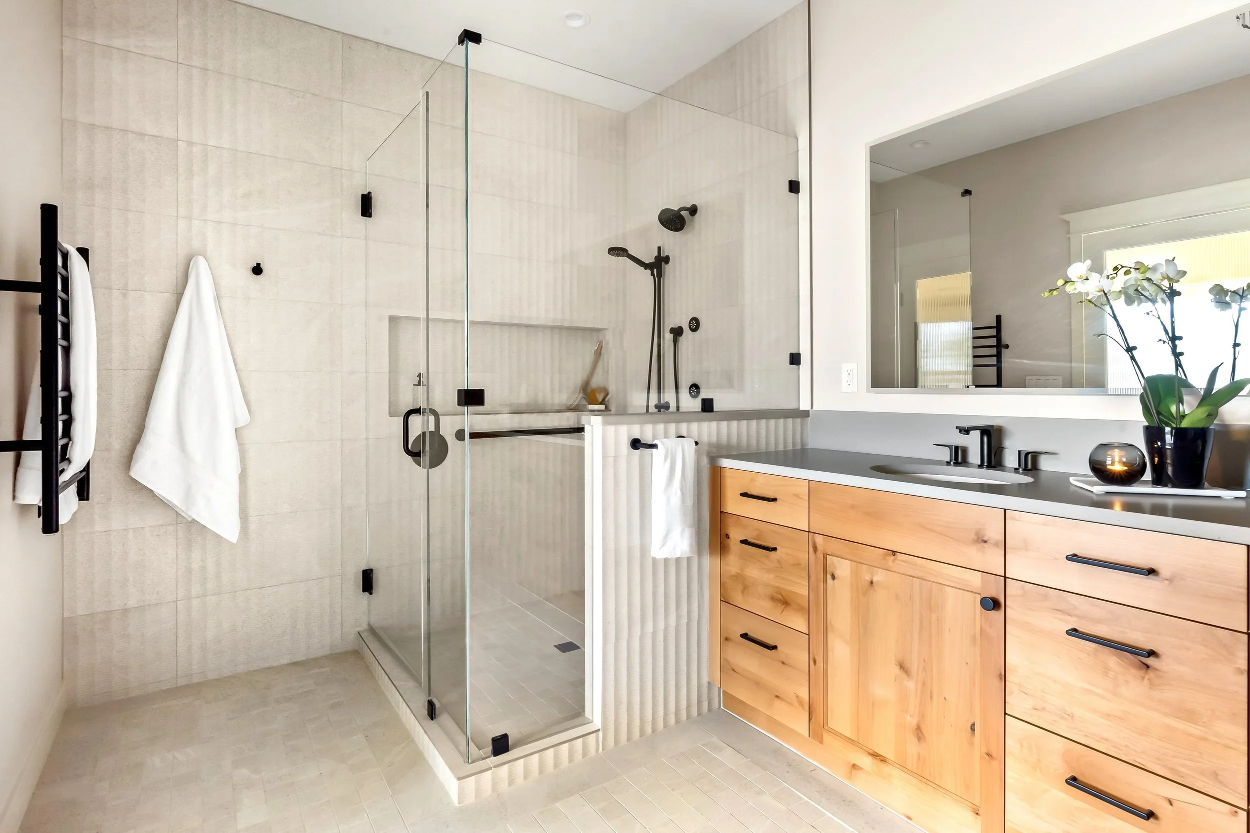 Modern bathroom with a glass shower enclosure, light-colored tiles, a wooden vanity with black handles, a large mirror, a potted plant, and a black candle holder.