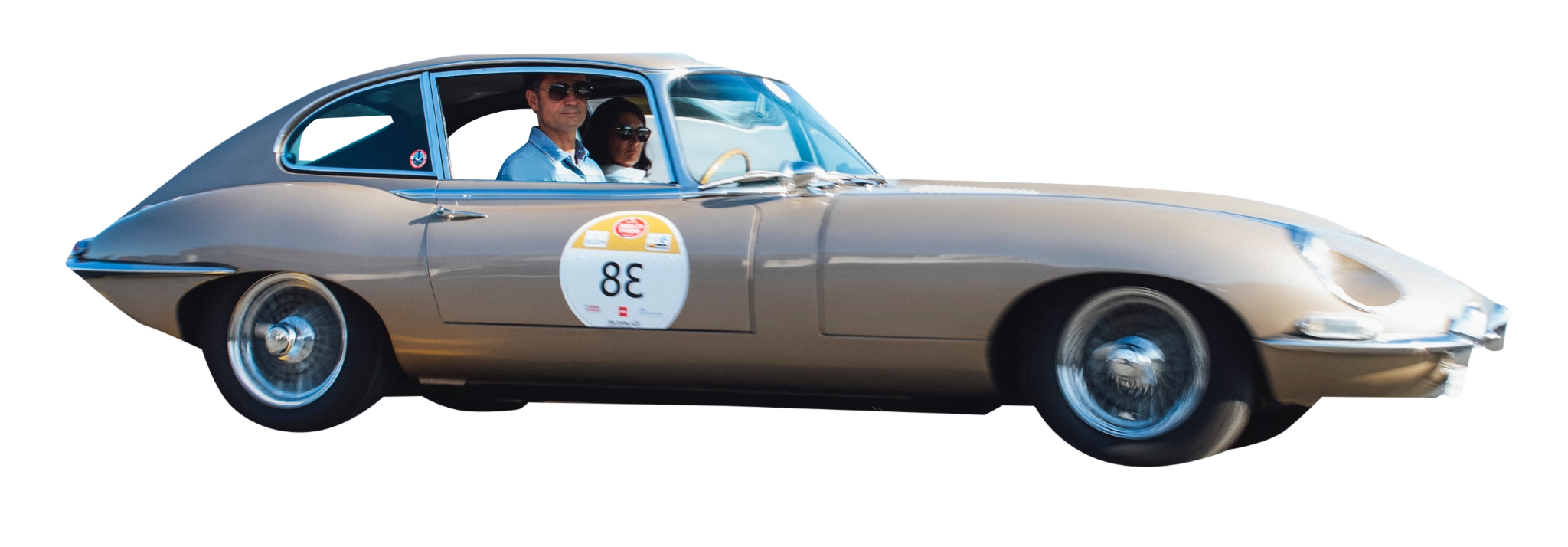 A vintage silver sports car with two people inside, both wearing sunglasses, participating in a racing event indicated by a round white sticker with the number 38 on the door.