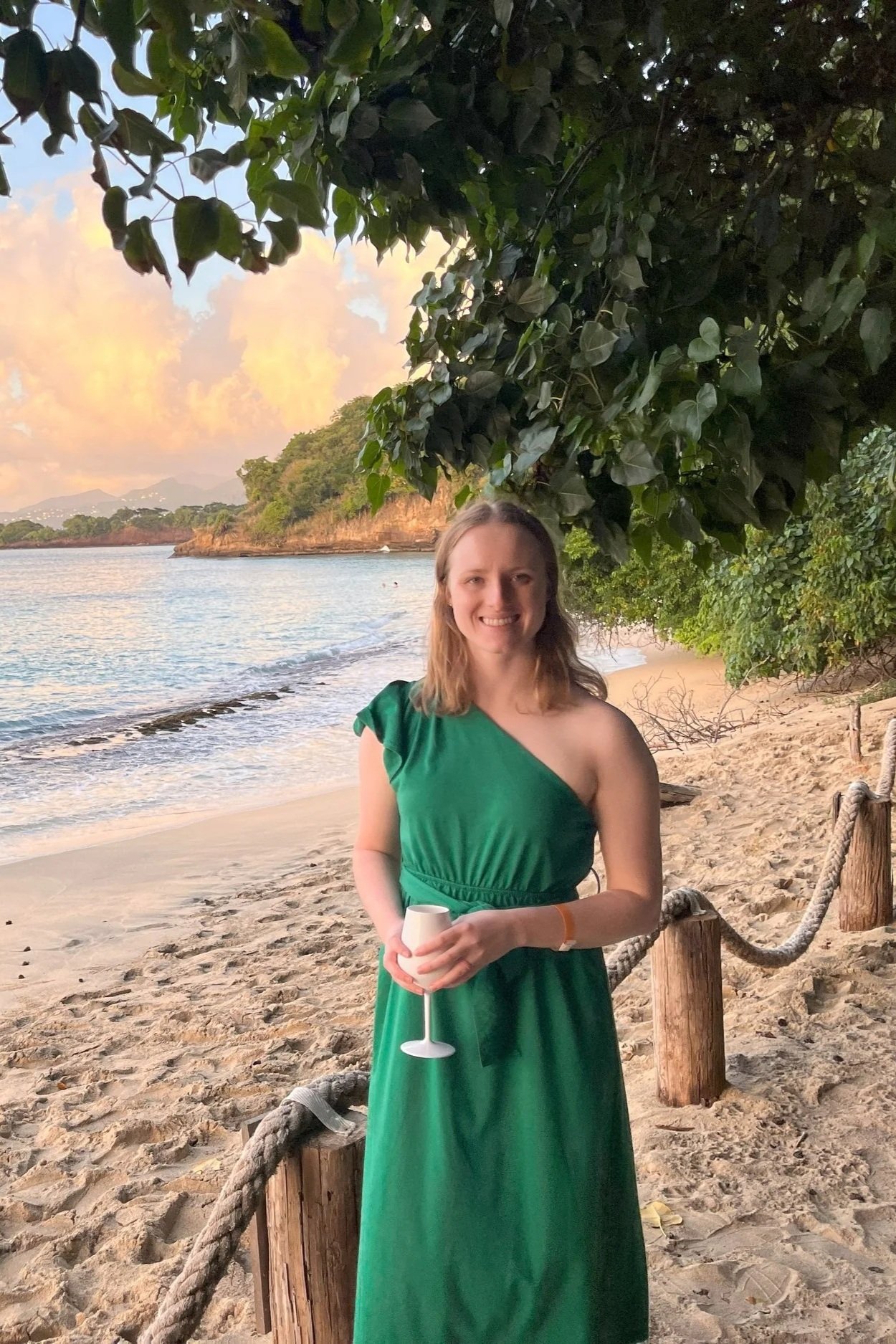 Rachel Fields in a green dress standing on a sandy beach, holding a white drink glass, smiling, with trees, water, and a colorful sky in the background.
