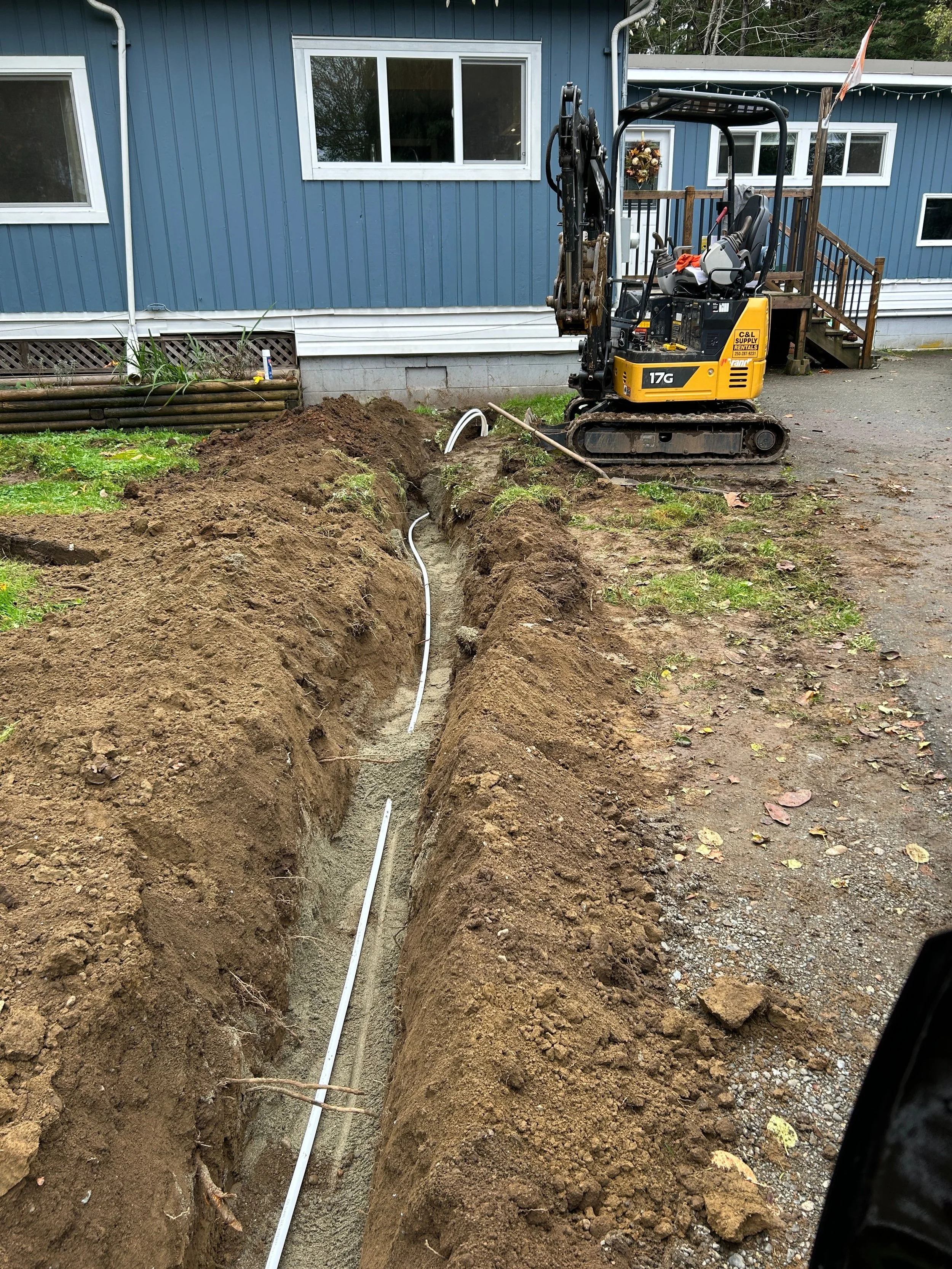 A construction site showing a narrow trench in the ground with a white pipe running through it. A small yellow excavator is parked nearby, and a blue house with white-trimmed windows and a porch is visible in the background.