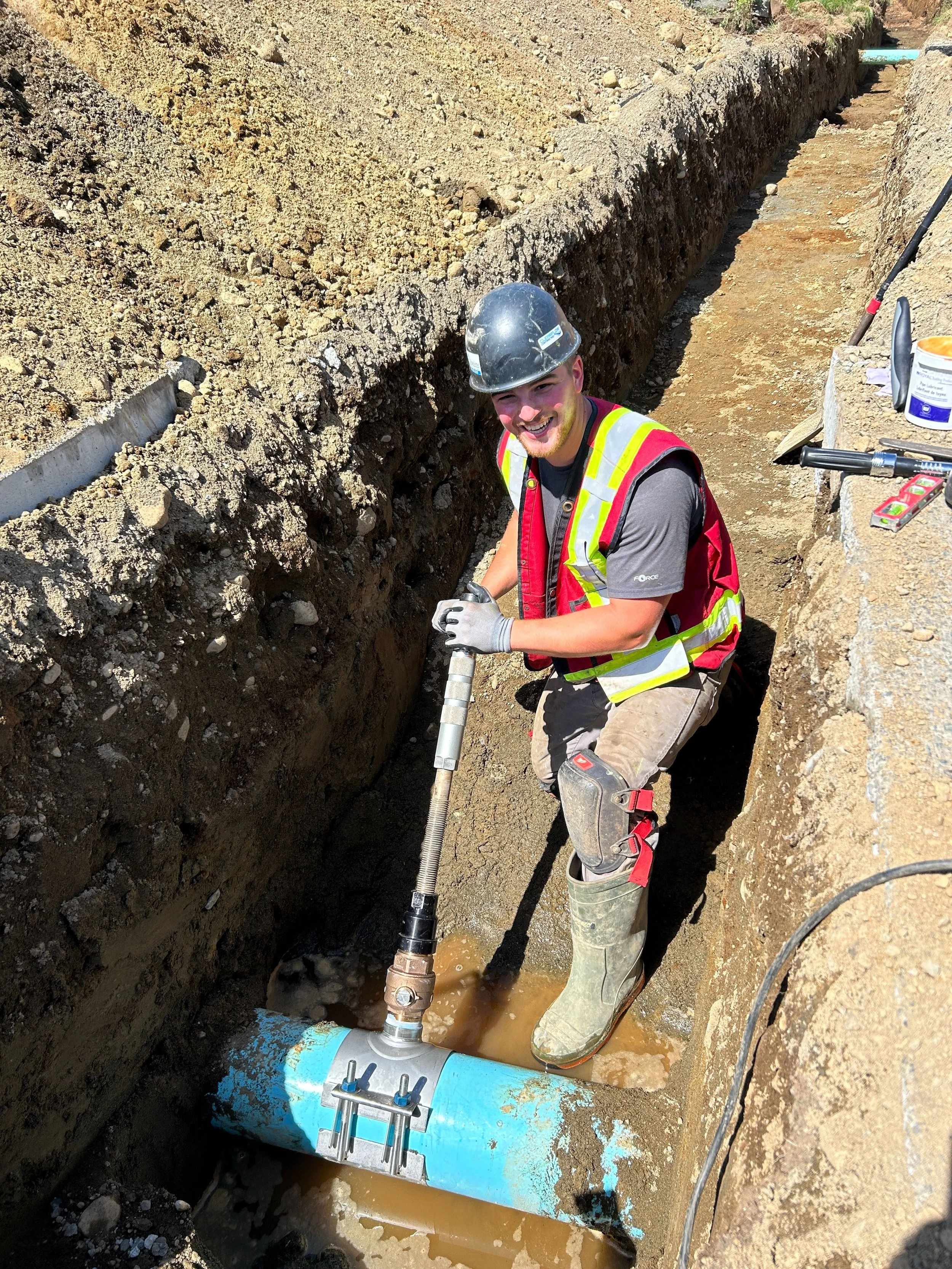 A construction worker wearing a hard hat, safety vest, and work boots is installing or repairing industrial piping in a trench at a construction site. The worker is smiling and holding a pipe fitting tool.