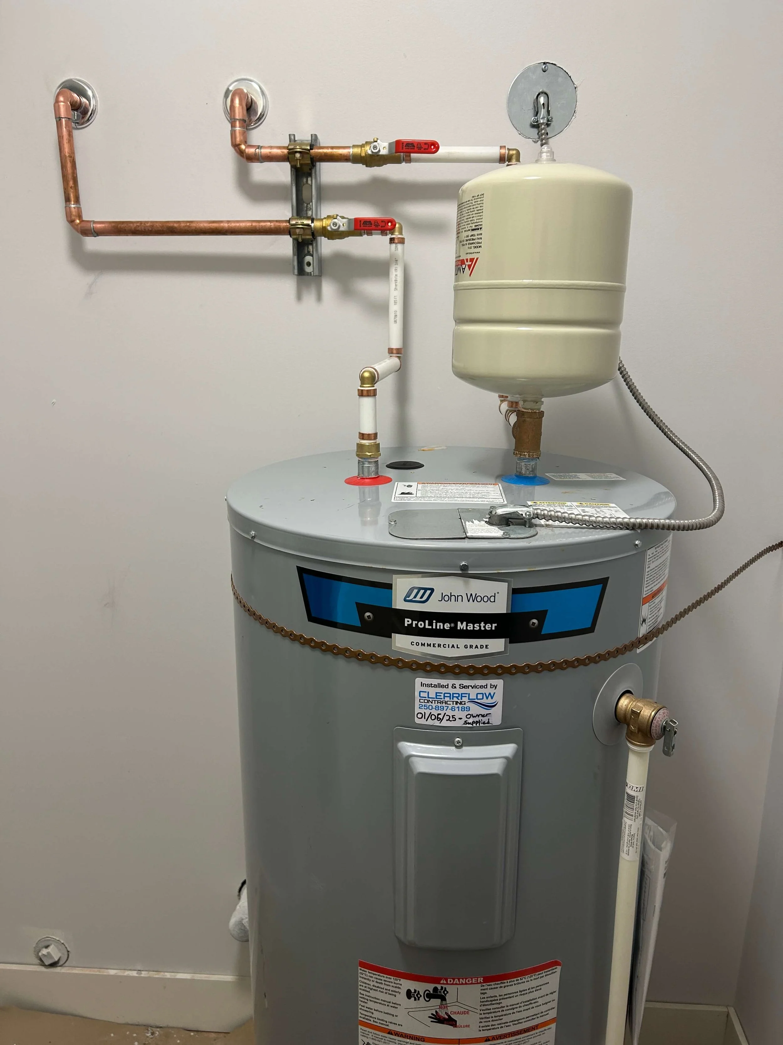 A commercial water heater with copper pipes and valves mounted on a beige wall, labeled 'John Wood ProLine Master' and installed by ClearFlow Contracting.