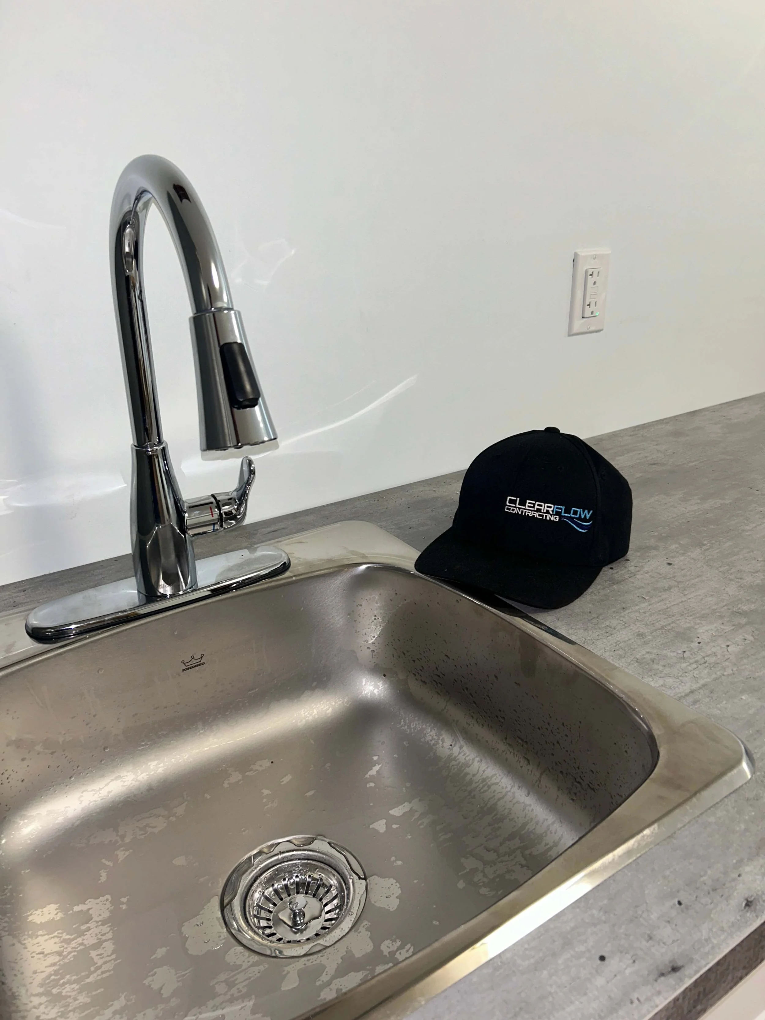 Kitchen stainless steel sink with a chrome faucet, and a black cap with the text 'Clear Flow Contracting' on it placed on the counter next to the sink. The background includes a white wall with an electrical outlet.
