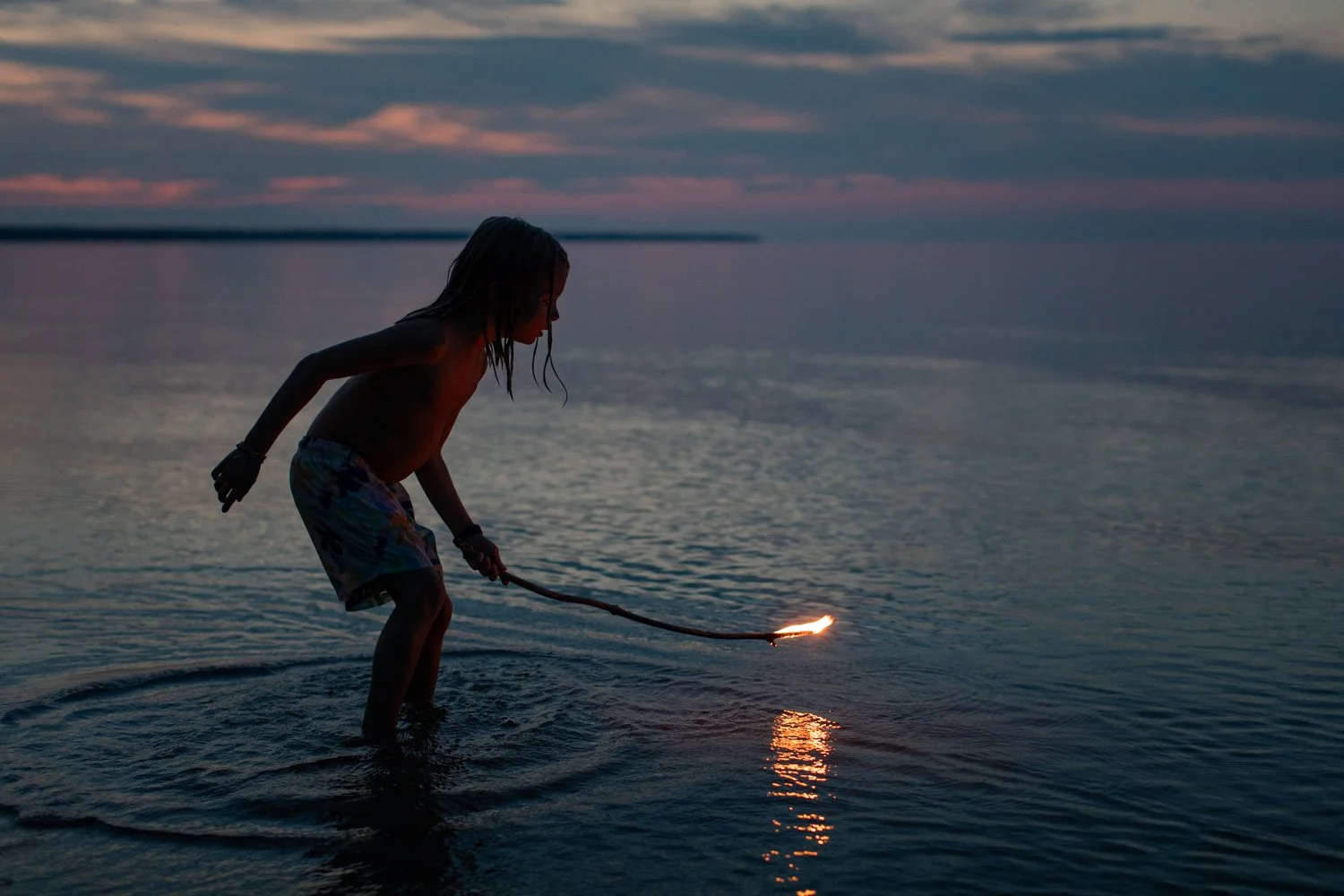 A child plays in shallow water at sunset while holding a small burning torch.