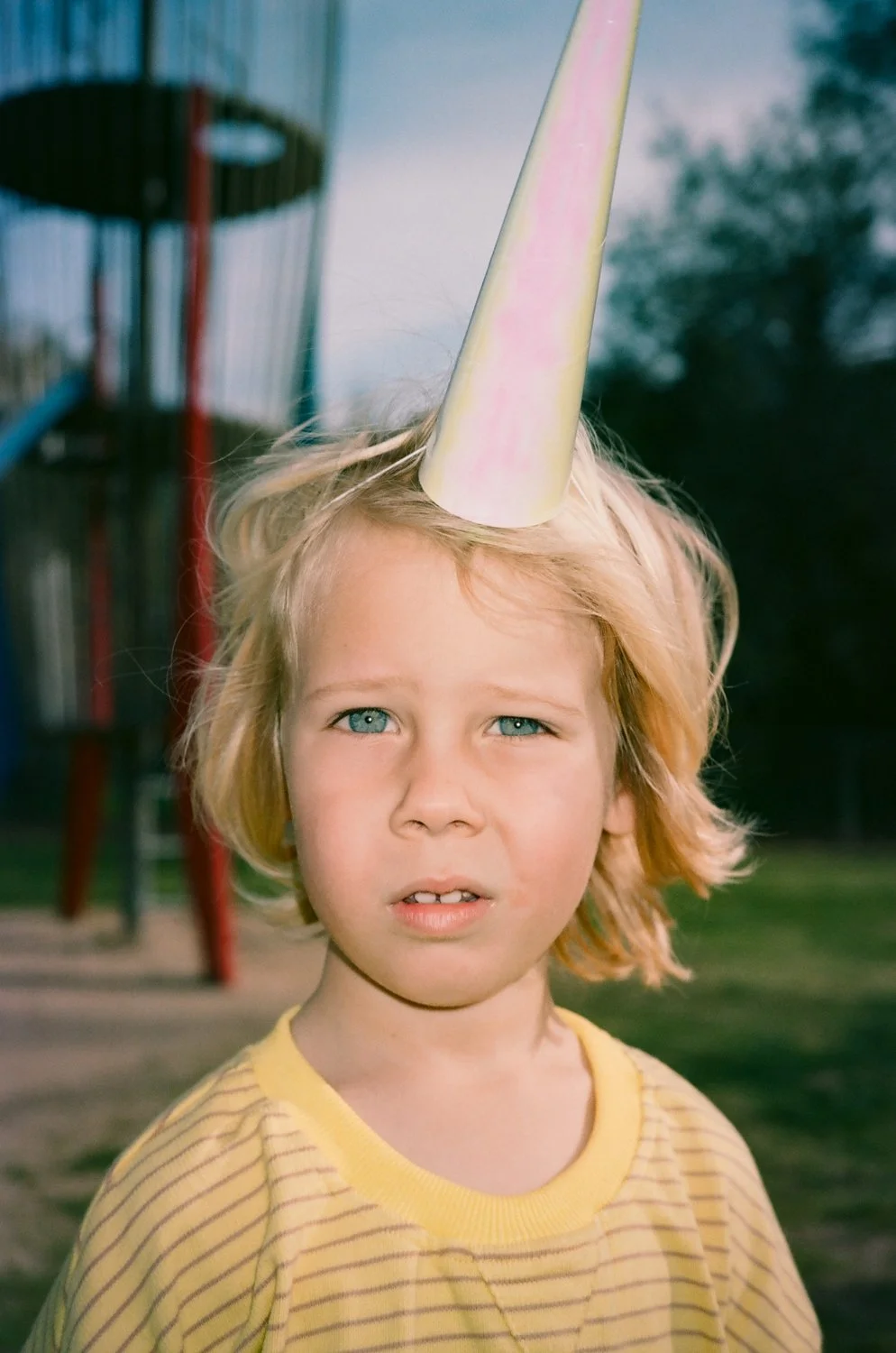 A child wearing a colorful party hat looks toward the camera at a playground.
