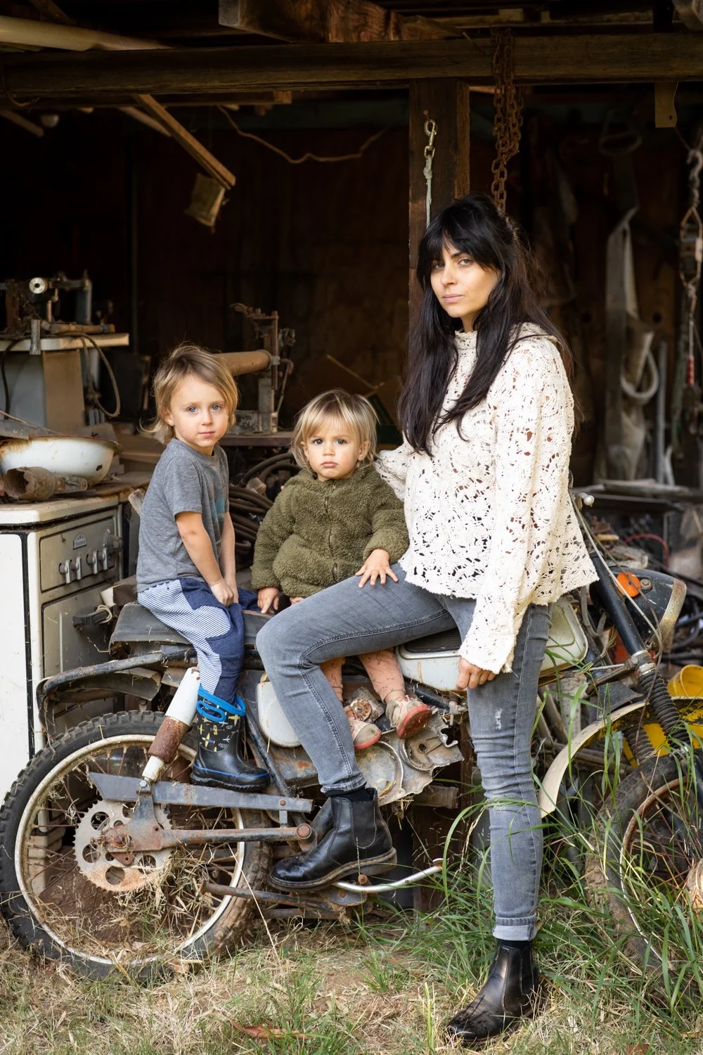 A mother sits with her two children on an old farm vehicle surrounded by tools and garden equipment.