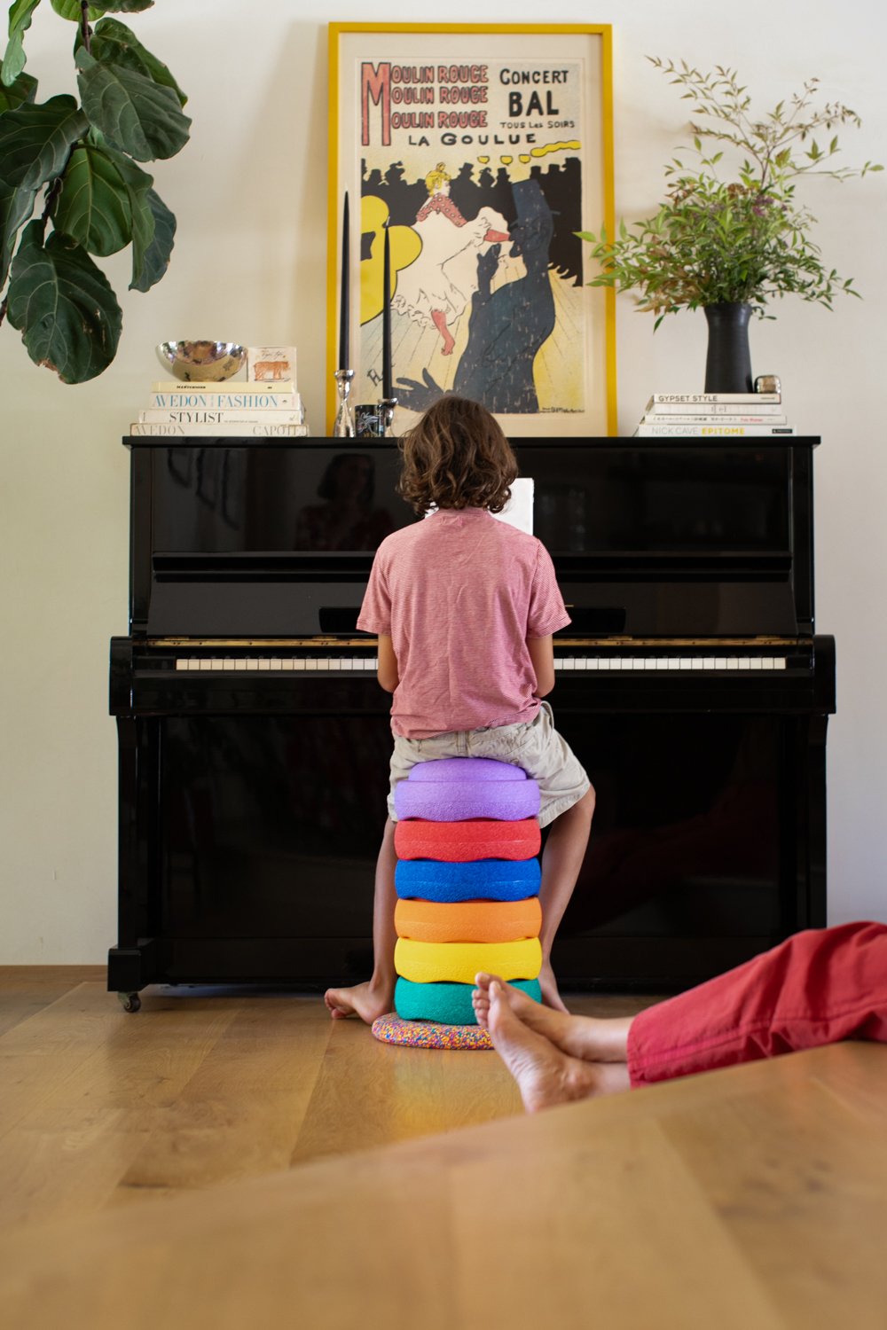 A child plays the piano while sitting on a stack of colorful cushions at home.