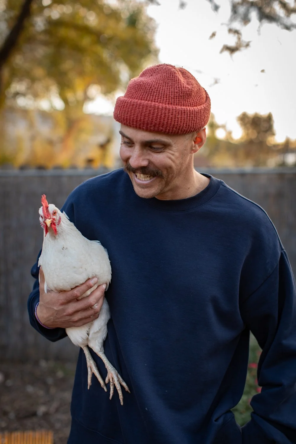 A man smiles while holding a chicken outdoors in his backyard.