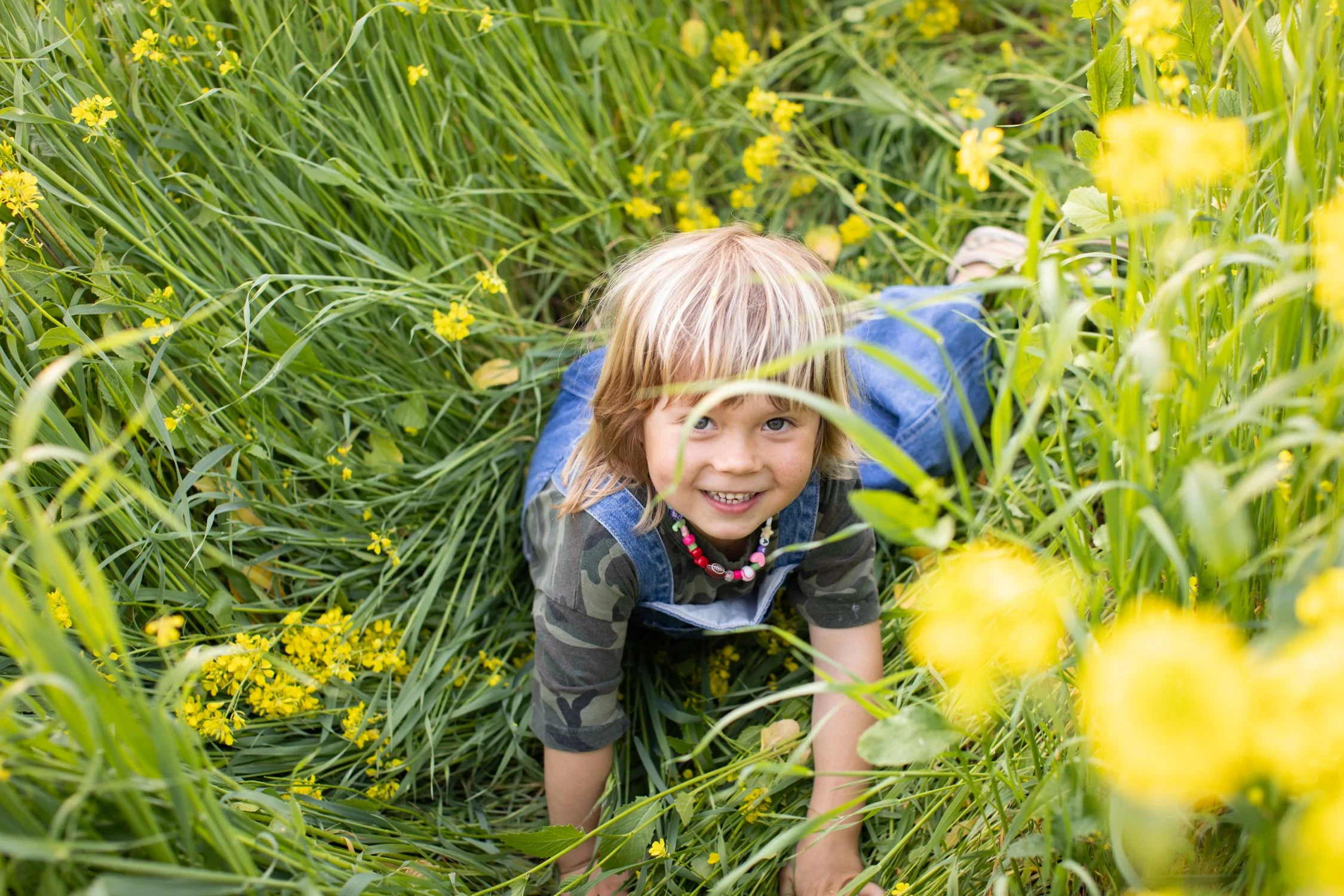 A smiling child crouches among tall grass and yellow flowers in the garden.