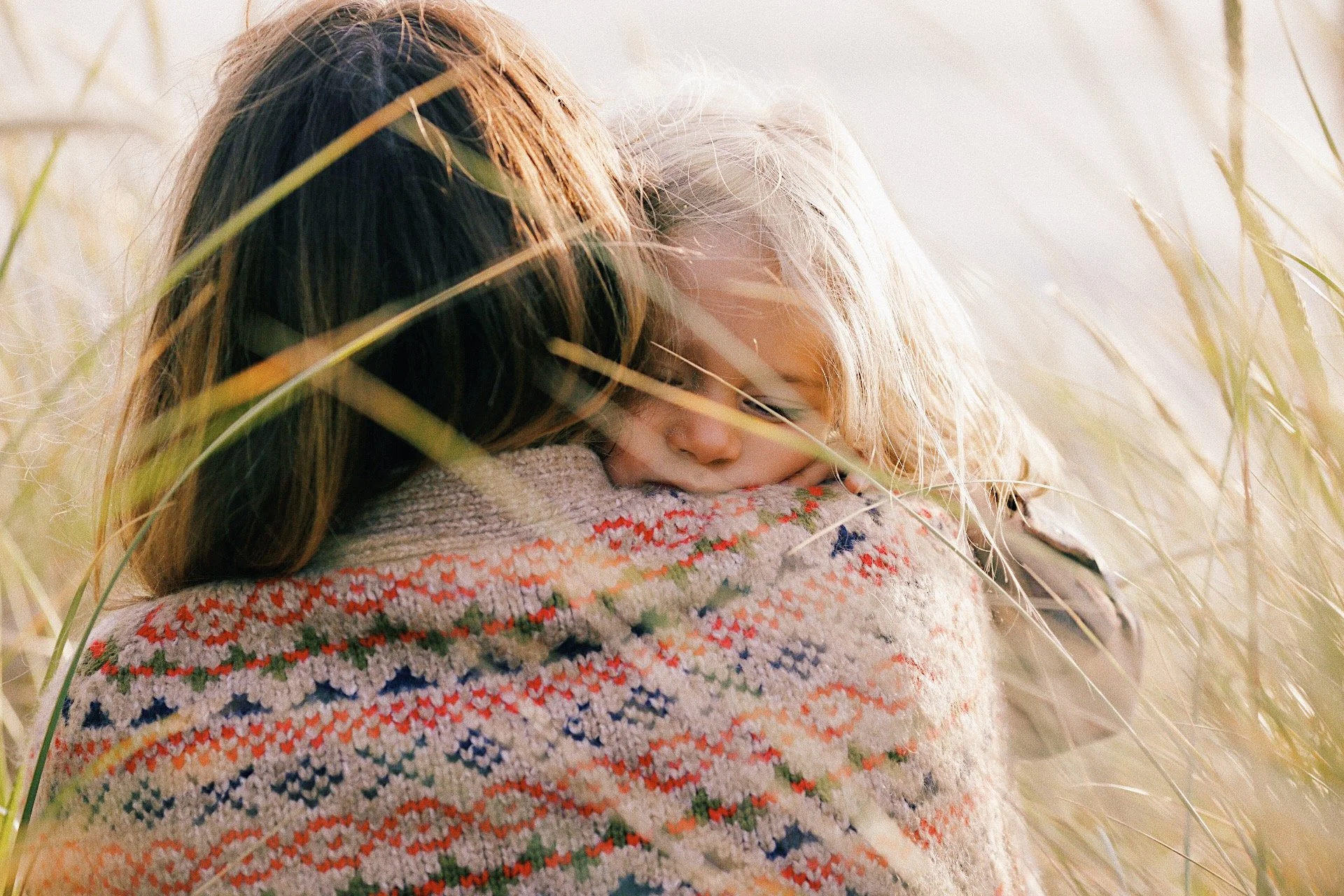A mother holds her child close while they sit together in tall grass.