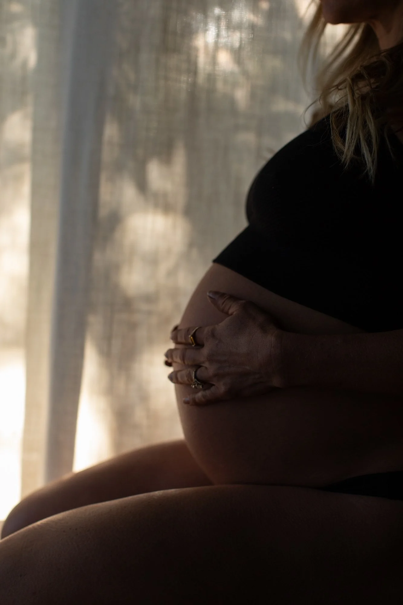 A pregnant woman rests her hands on her belly in soft window light.