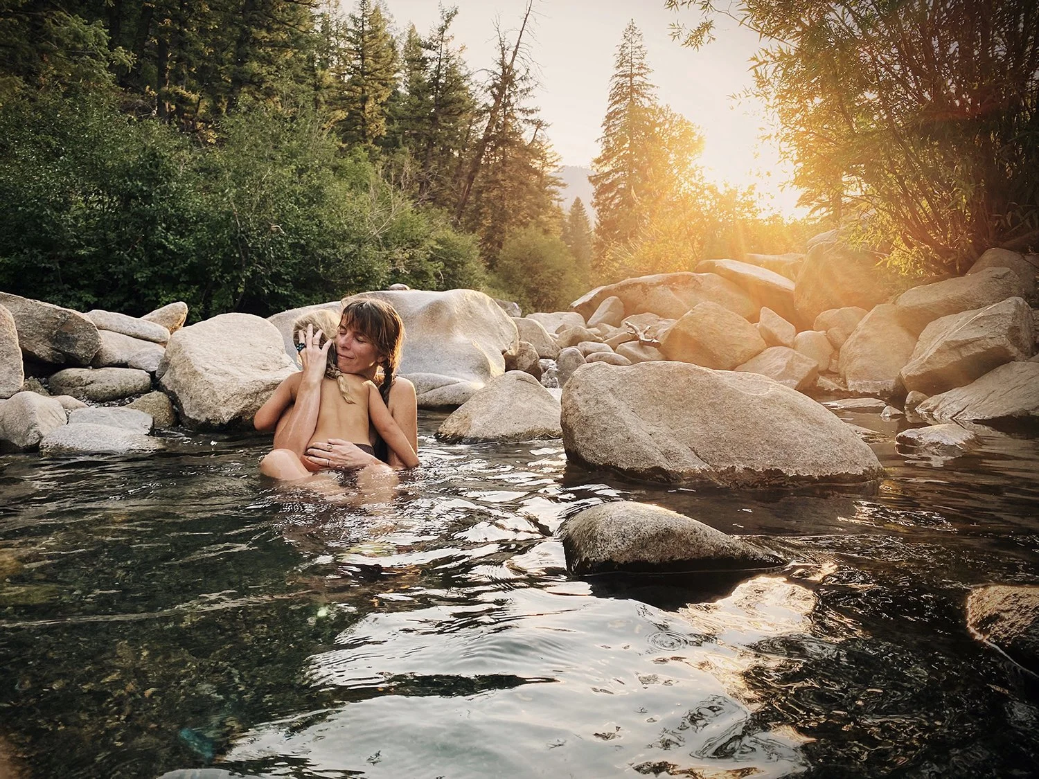 A child sits in a mountain stream surrounded by rocks and trees in the warm afternoon light.
