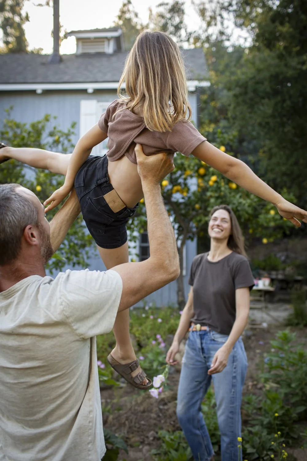 A father lifts his daughter playfully in the garden while her mother watches and smiles.