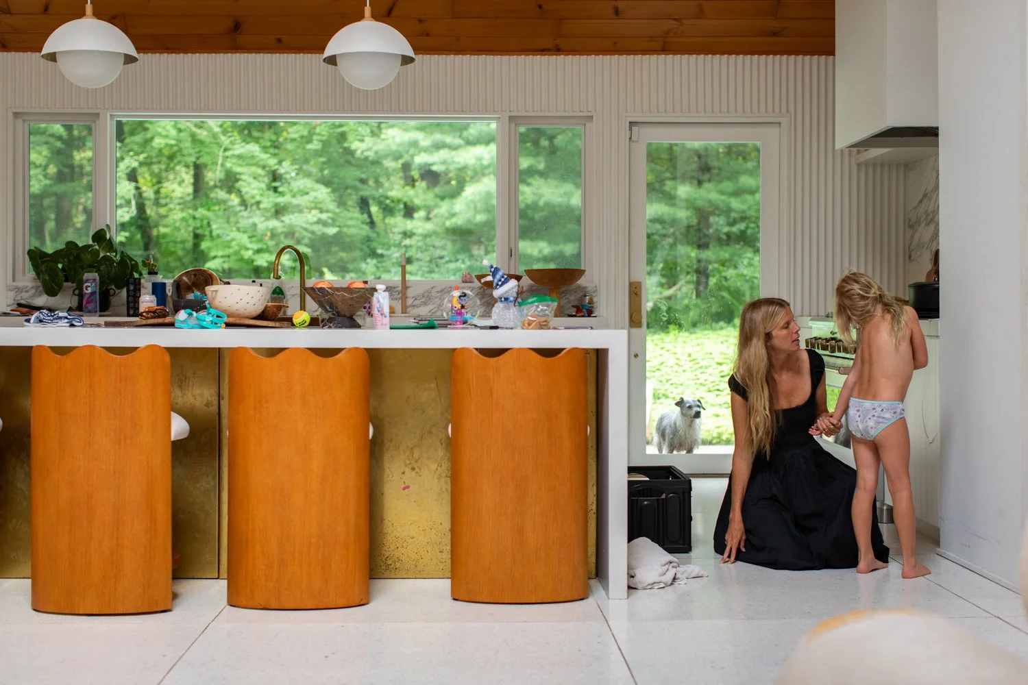 A mother helps her daughter get dressed in the kitchen while their dog watches nearby.