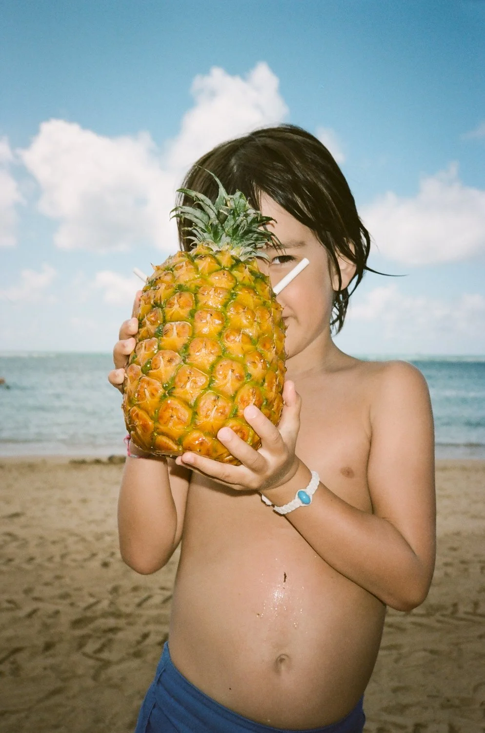 A child holds a pineapple in front of their face on a sunny beach.