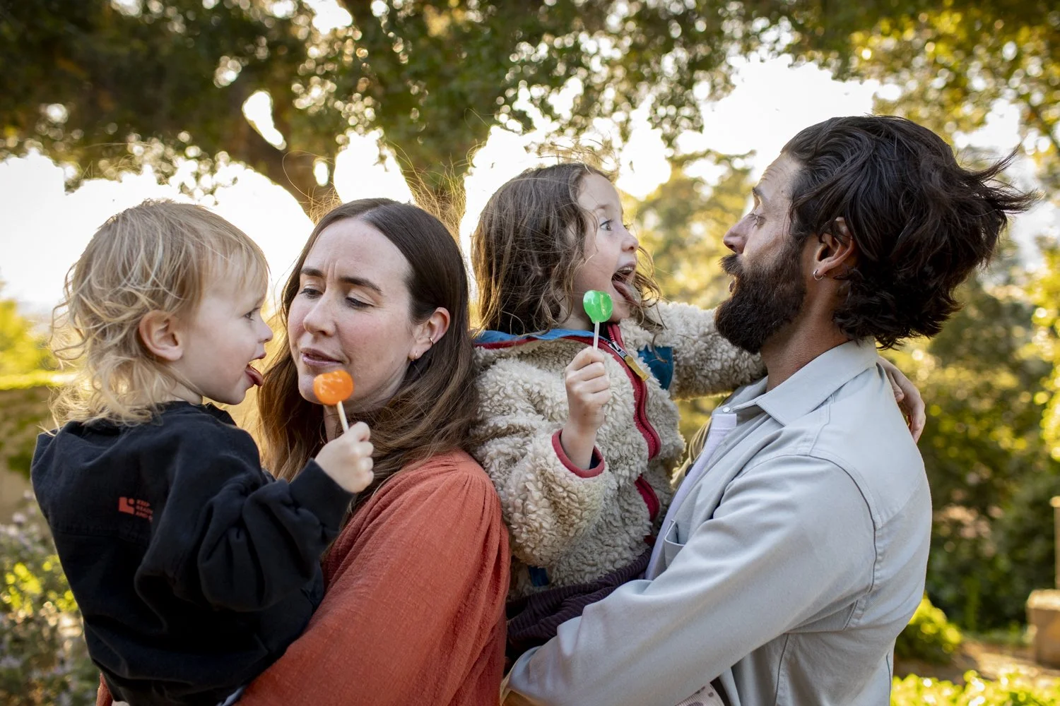A family shares a playful moment together outdoors, enjoying lollipops in the warm afternoon light.