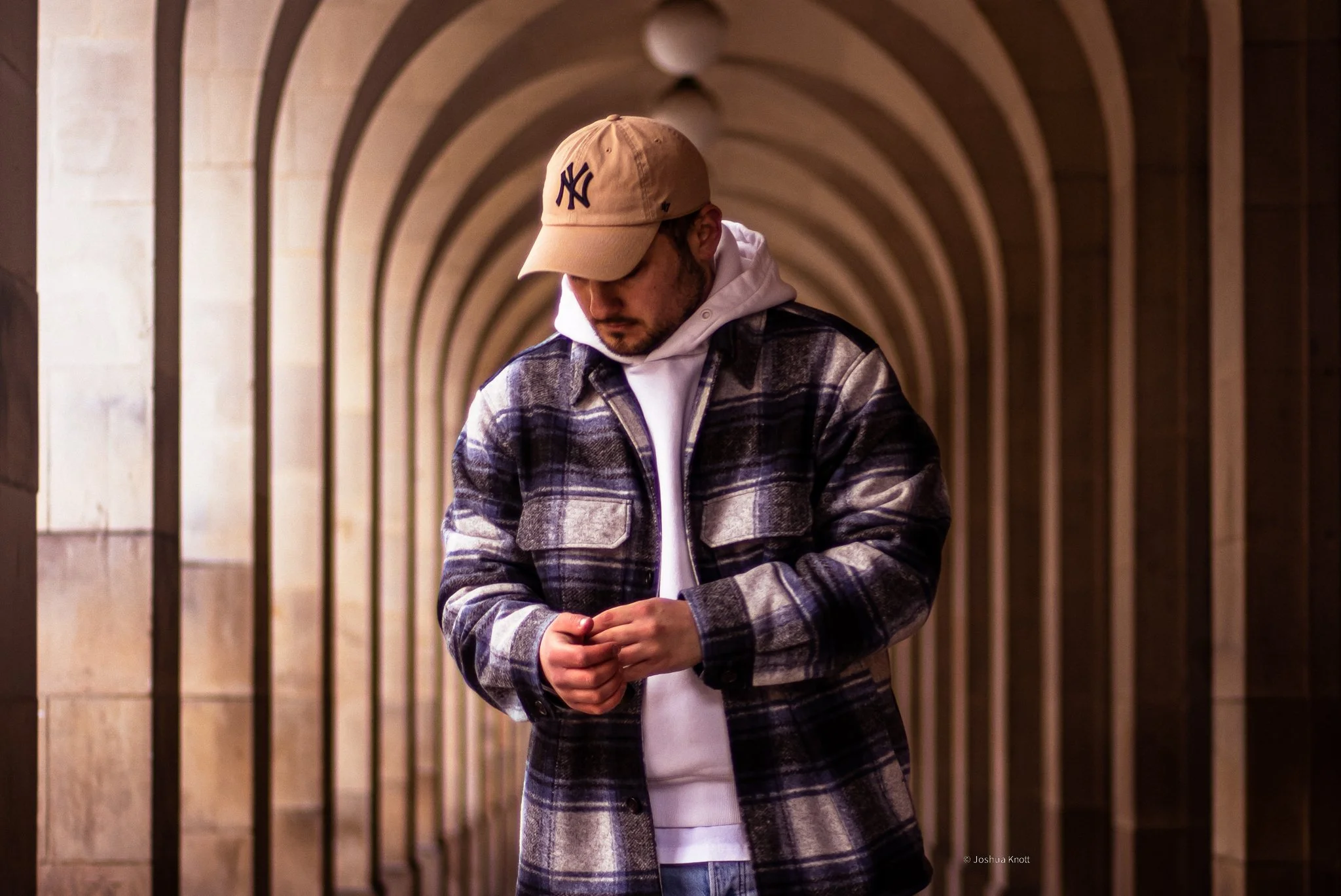Person wearing a plaid jacket, white hoodie, and beige cap in an arched corridor.