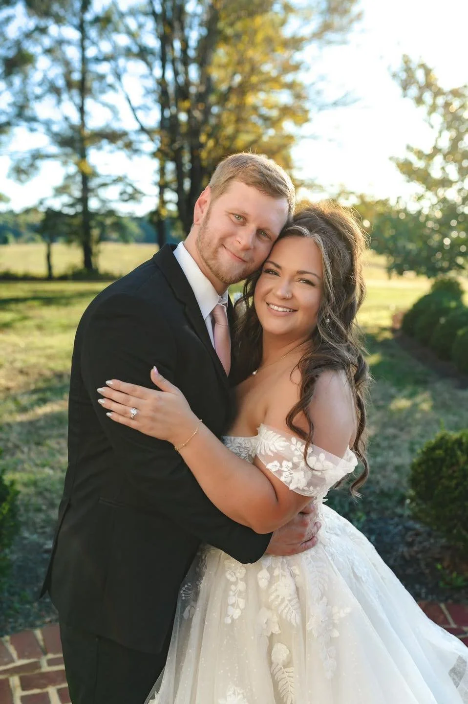 A newlywed couple hugging outdoors during sunset, with trees and a grassy field in the background.