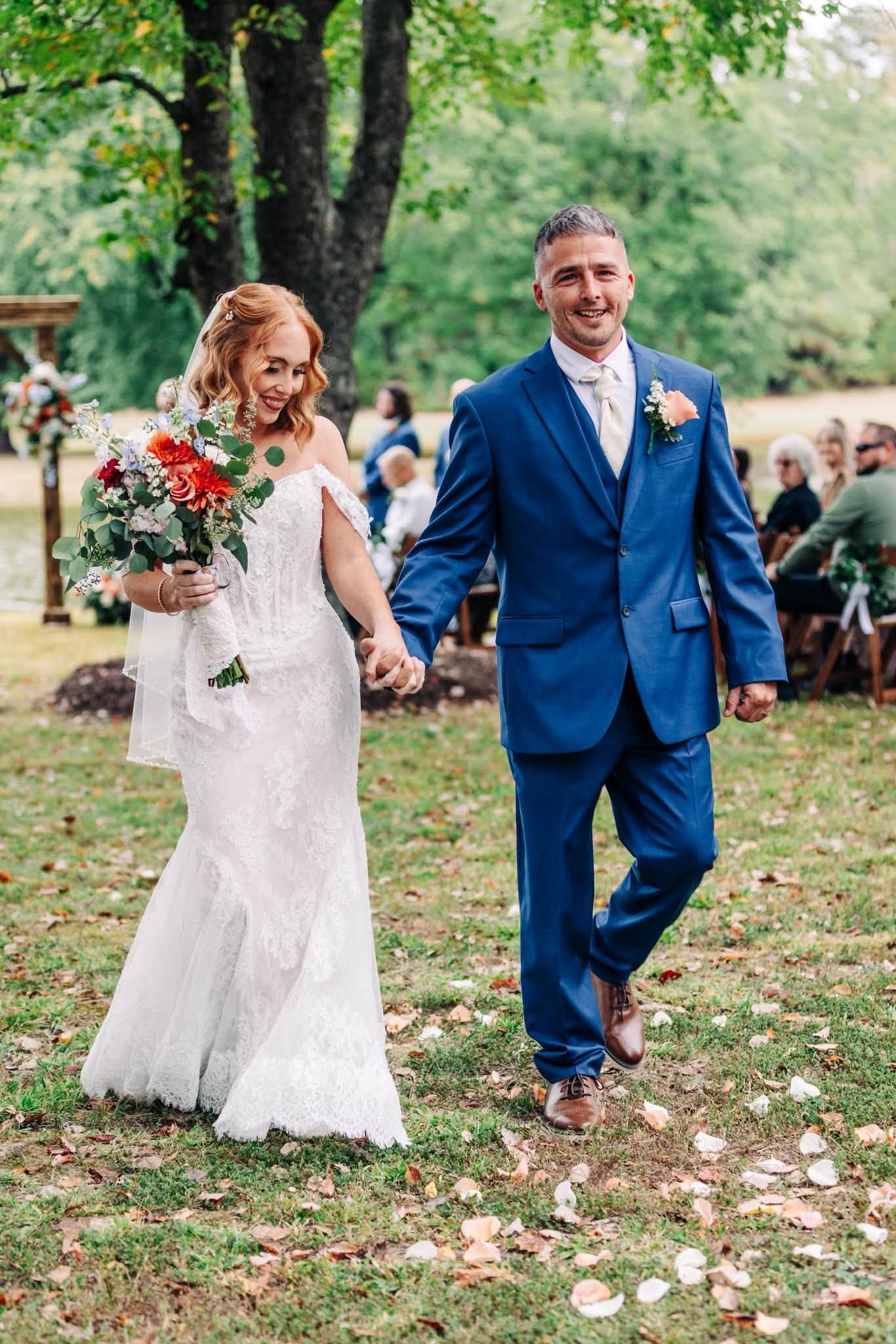A bride and groom walking hand in hand outdoors at a wedding ceremony, surrounded by greenery and guests seated in the background.