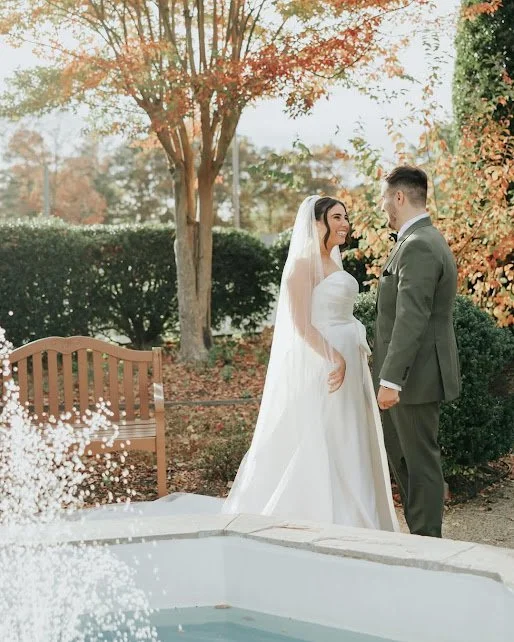 Bride and groom standing outdoors in a garden with autumn foliage, looking at each other, with a wooden bench and a fountain in the foreground.