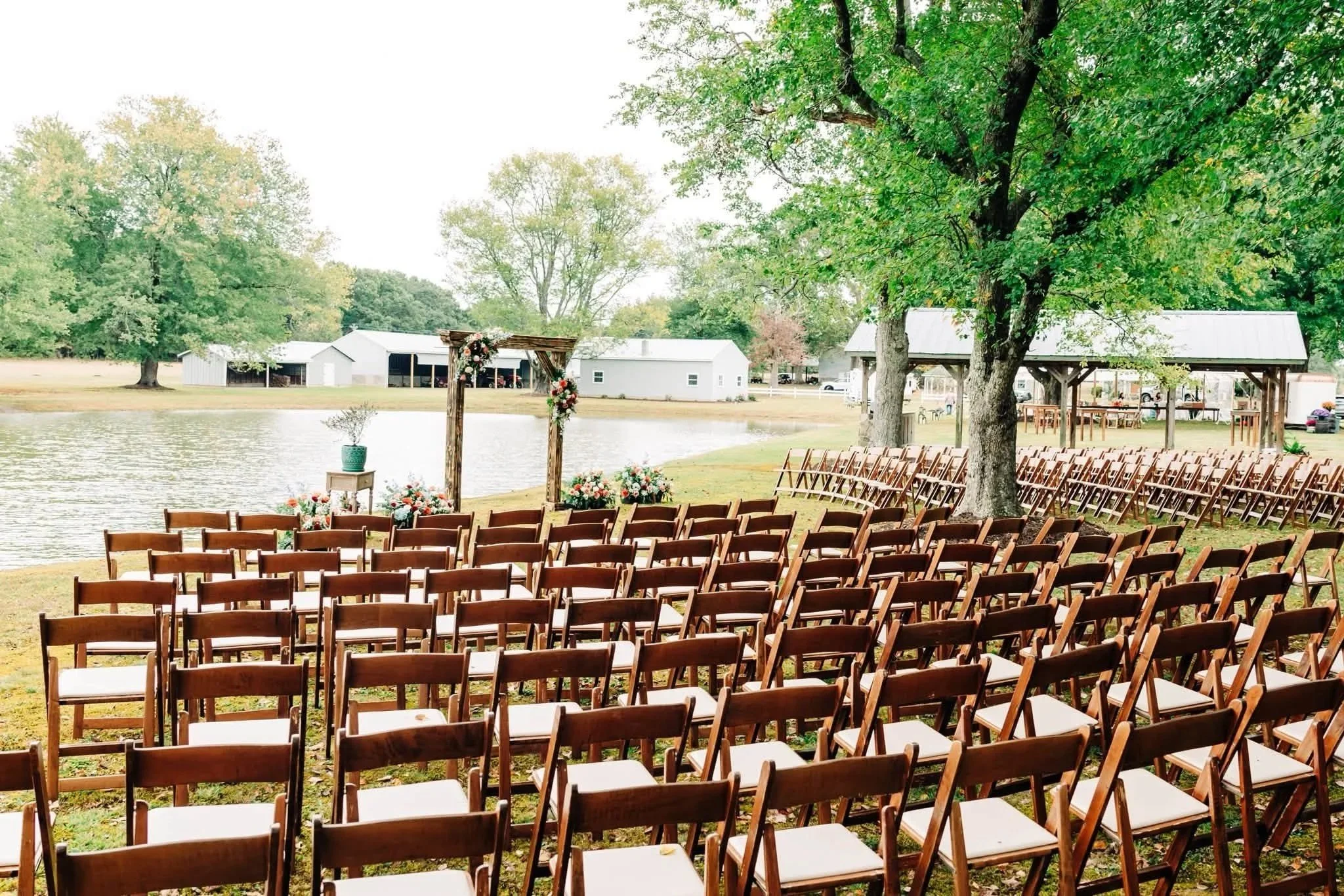 Outdoor wedding seating setup by a lake with rows of wooden chairs, a decorated arch, and trees in the background.