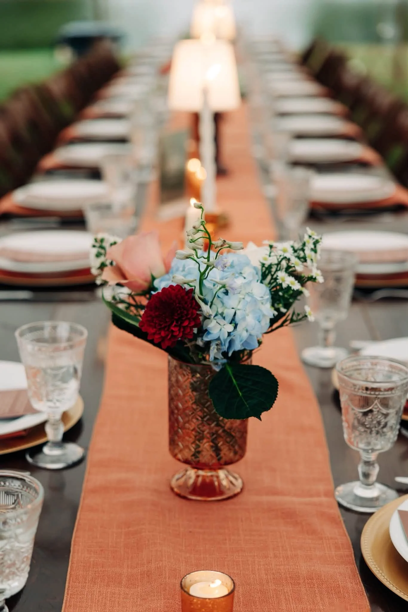 A long dining table with a burnt orange table runner, decorated with a floral centerpiece in a copper-colored vase. The table is set with white plates, water glasses, and candles in small holders, with a blurred outdoor background.