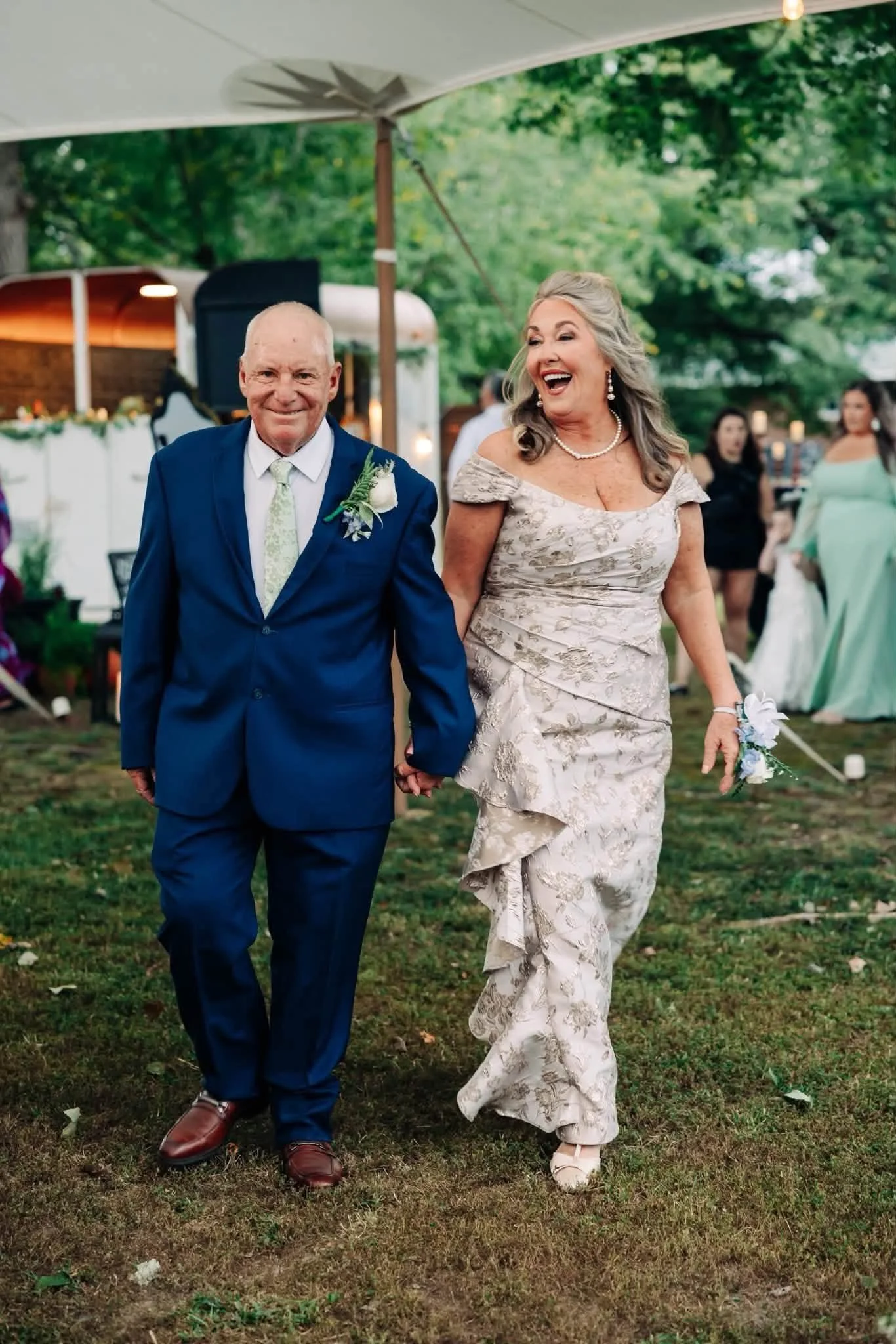 An elderly couple celebrating at a wedding reception outdoors, holding hands and smiling. The man wears a blue suit with a white shirt and a pale green tie, and the woman wears an off-shoulder, floral-patterned light-colored gown. They are walking on grass with trees and people in the background.