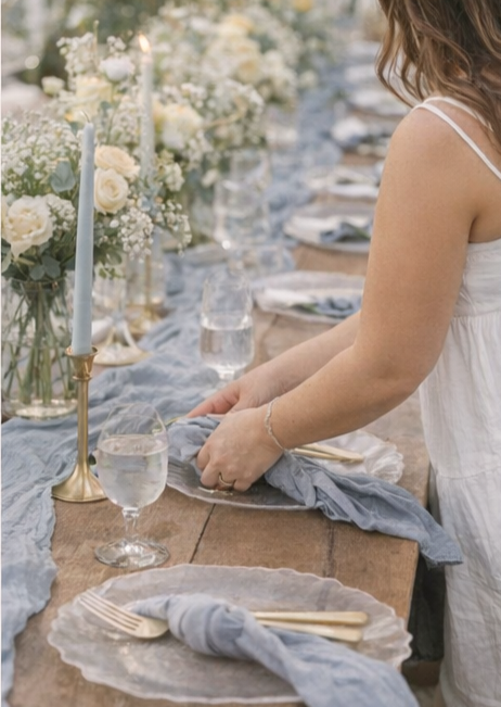 A woman adjusting place settings at a wooden table with blue accents and fresh florals