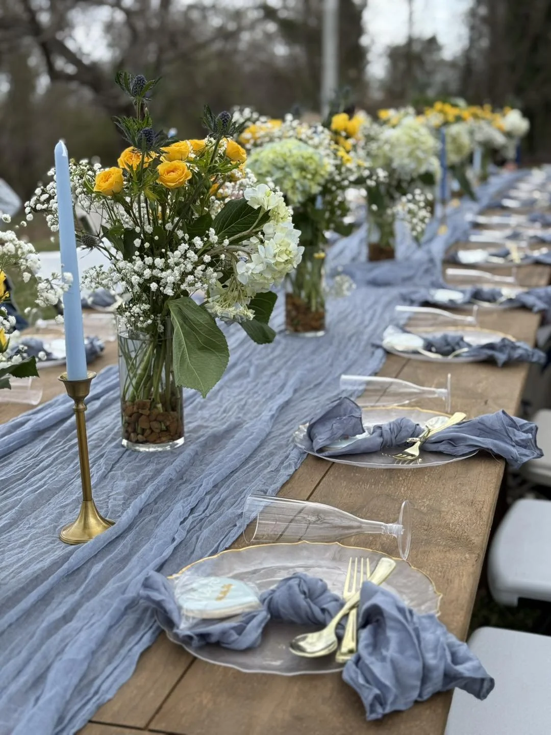 A long outdoor dining table decorated with a blue table runner, floral centerpieces with yellow and white flowers, and place settings with gold utensils and grey napkins tied with golden rings, set for a celebration.