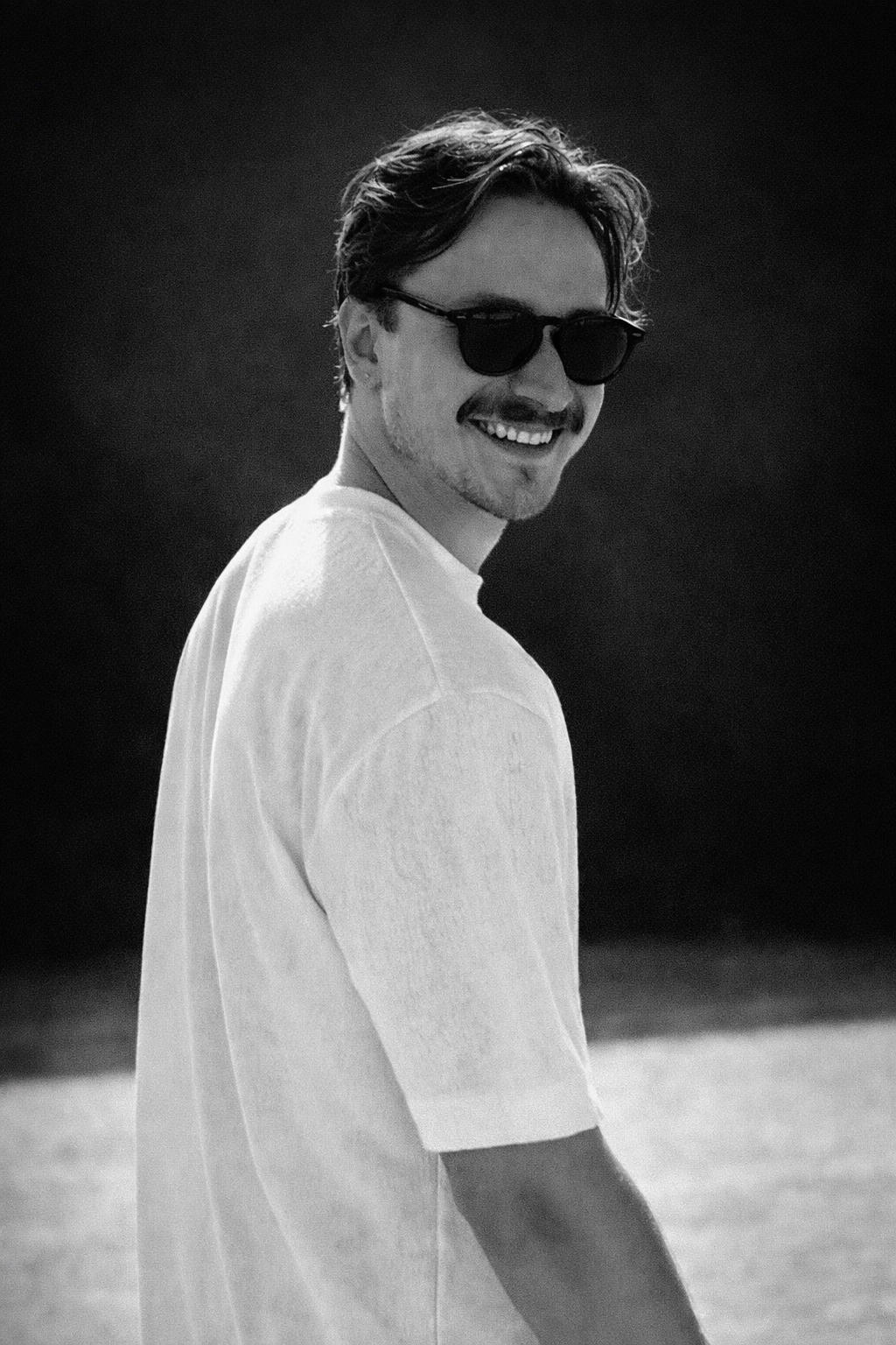 Camu Creutz wearing sunglasses and a plain white t-shirt, smiling and looking over his shoulder outdoors.