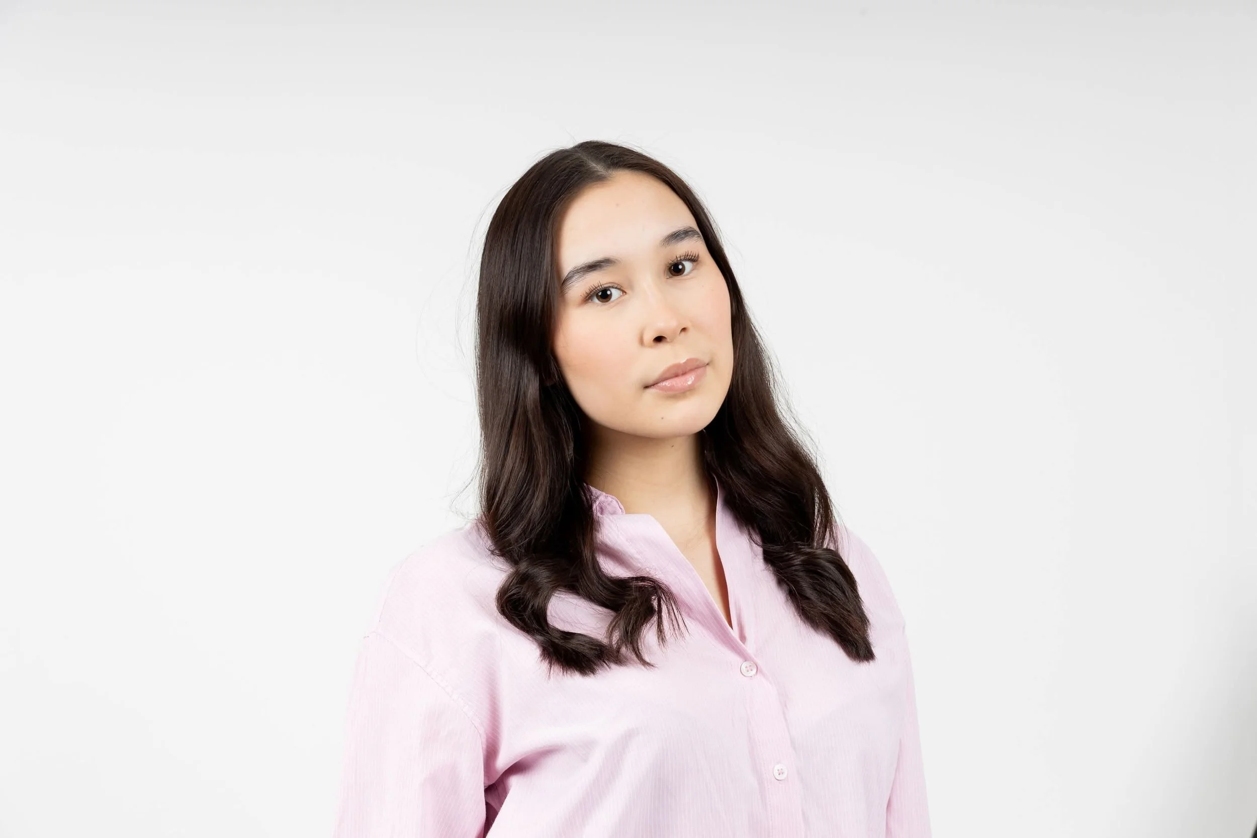 A young woman with long dark hair wearing a light pink button-up shirt against a white background.