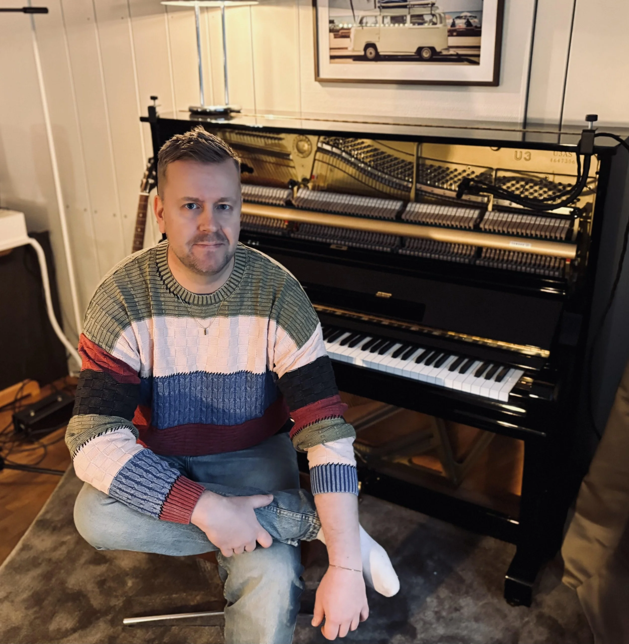 A man with short hair and a beard sitting on a stool in front of an upright piano with its lid open, revealing the strings and hammers, in a room with wood-paneled walls and a framed picture of a van hanging above the piano.