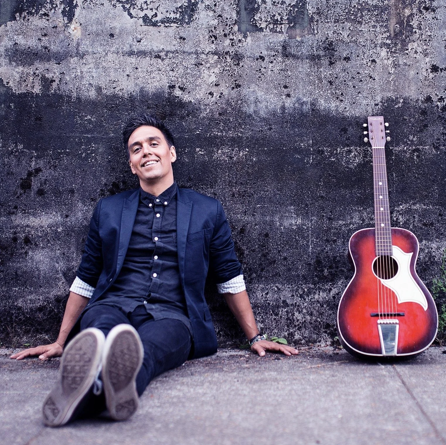 A young man in a dark suit sitting on the ground, leaning against a weathered wall, with a red acoustic guitar resting on the ground beside him.