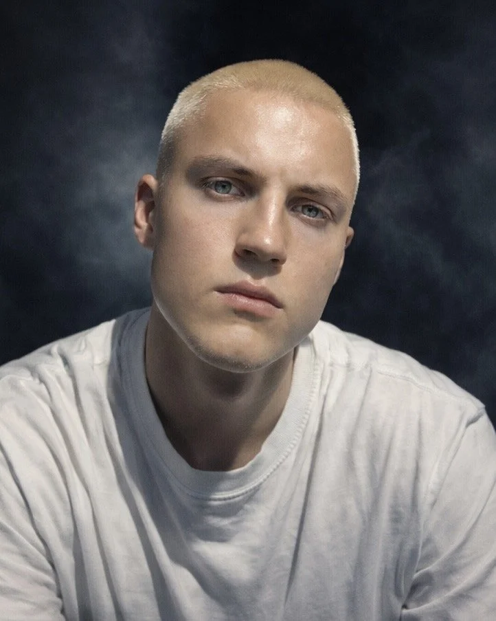 Elias Hjelm with a serious expression, light skin, short blond Hair, and wearing a white T-shirt, poses against a dark, textured backdrop.