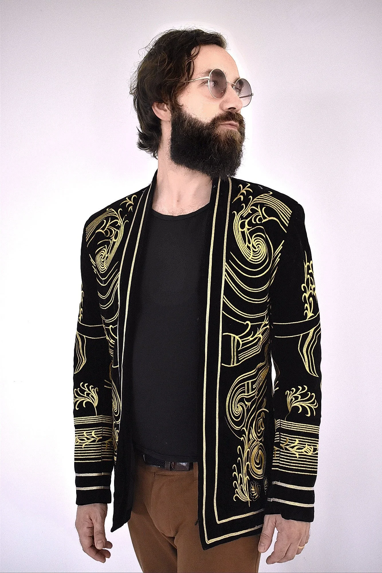 A man with long dark hair and a full beard wearing round sunglasses, a black shirt, and a black jacket with intricate gold embroidery, standing against a plain white background.