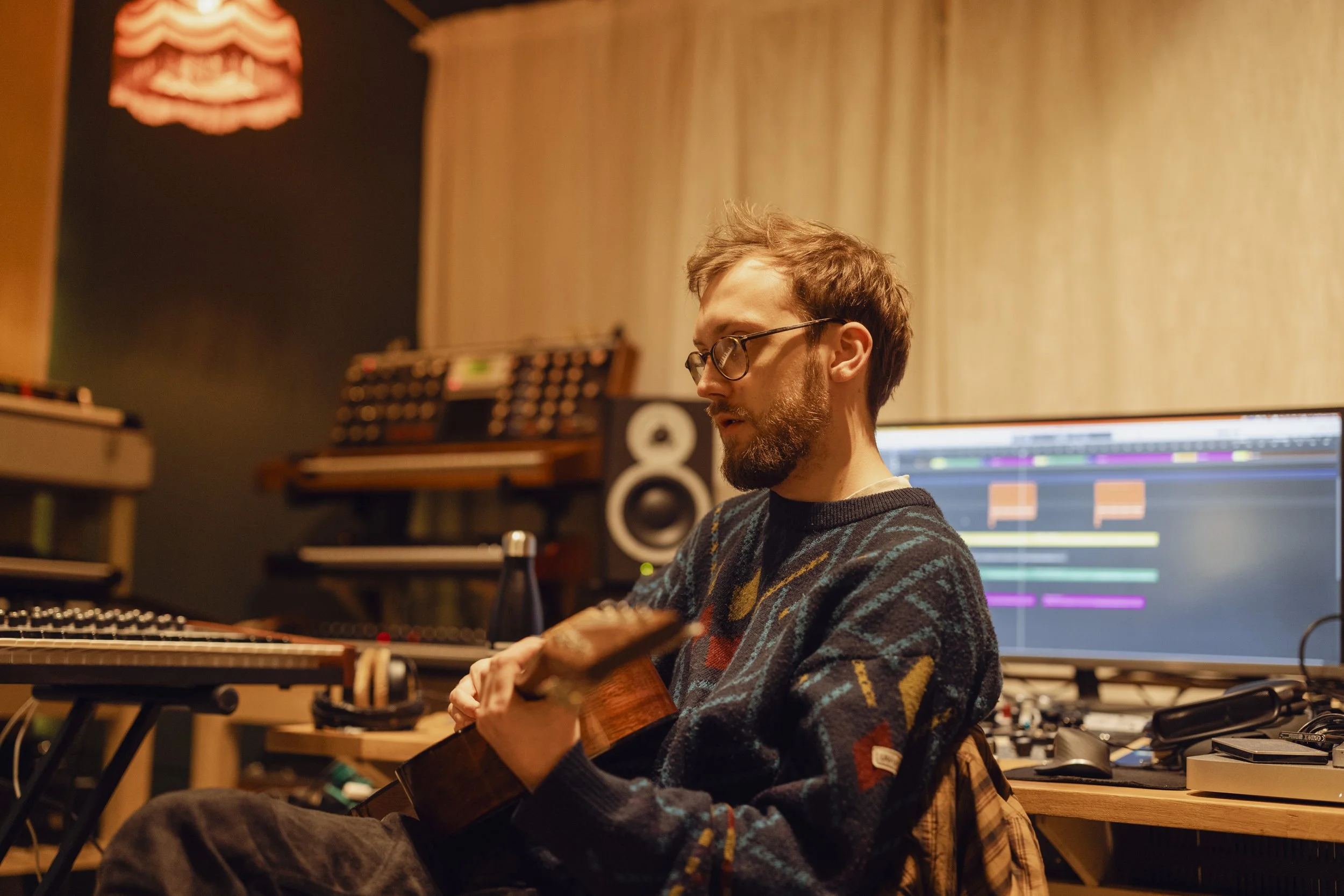 A man with glasses and a beard playing an acoustic guitar in a music studio, with a computer screen displaying music editing software in the background.