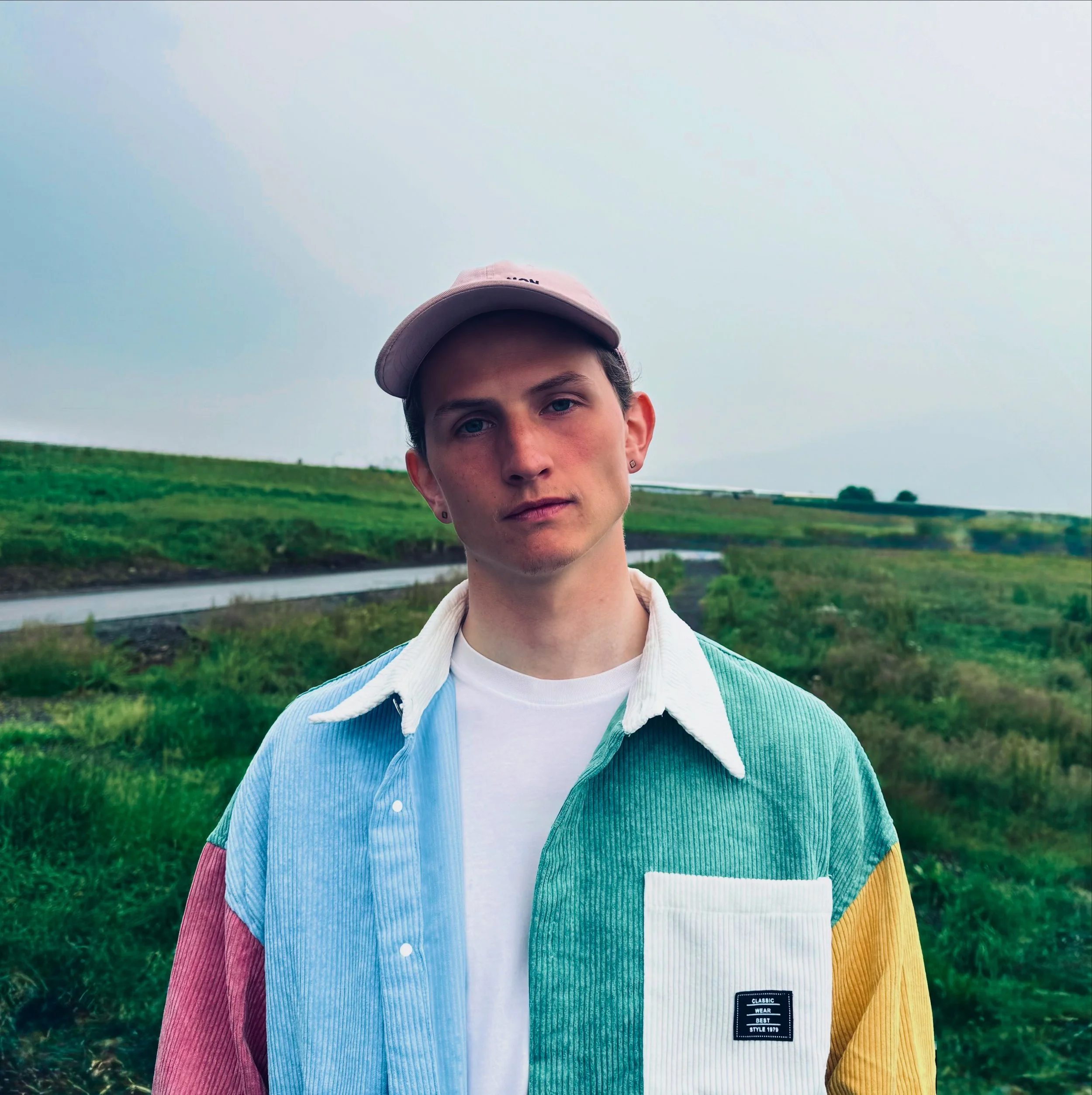 Atli Stefánsson standing outdoors in a green landscape with fields and a water body in the background. He is wearing a colorful patchwork shirt, a white t-shirt, and a pink cap.