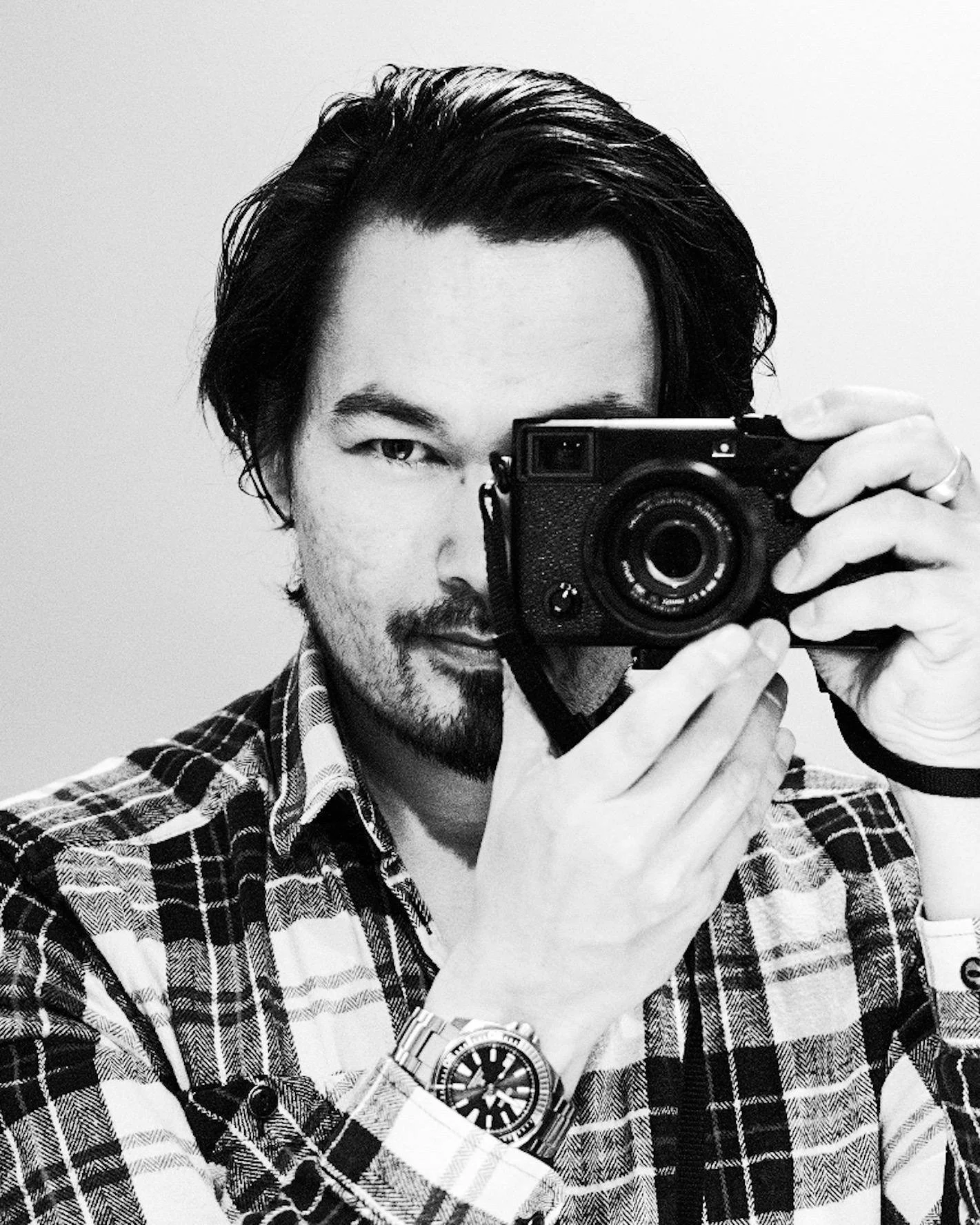 A man taking a selfie in front of a mirror with a camera, wearing a plaid shirt and a watch.