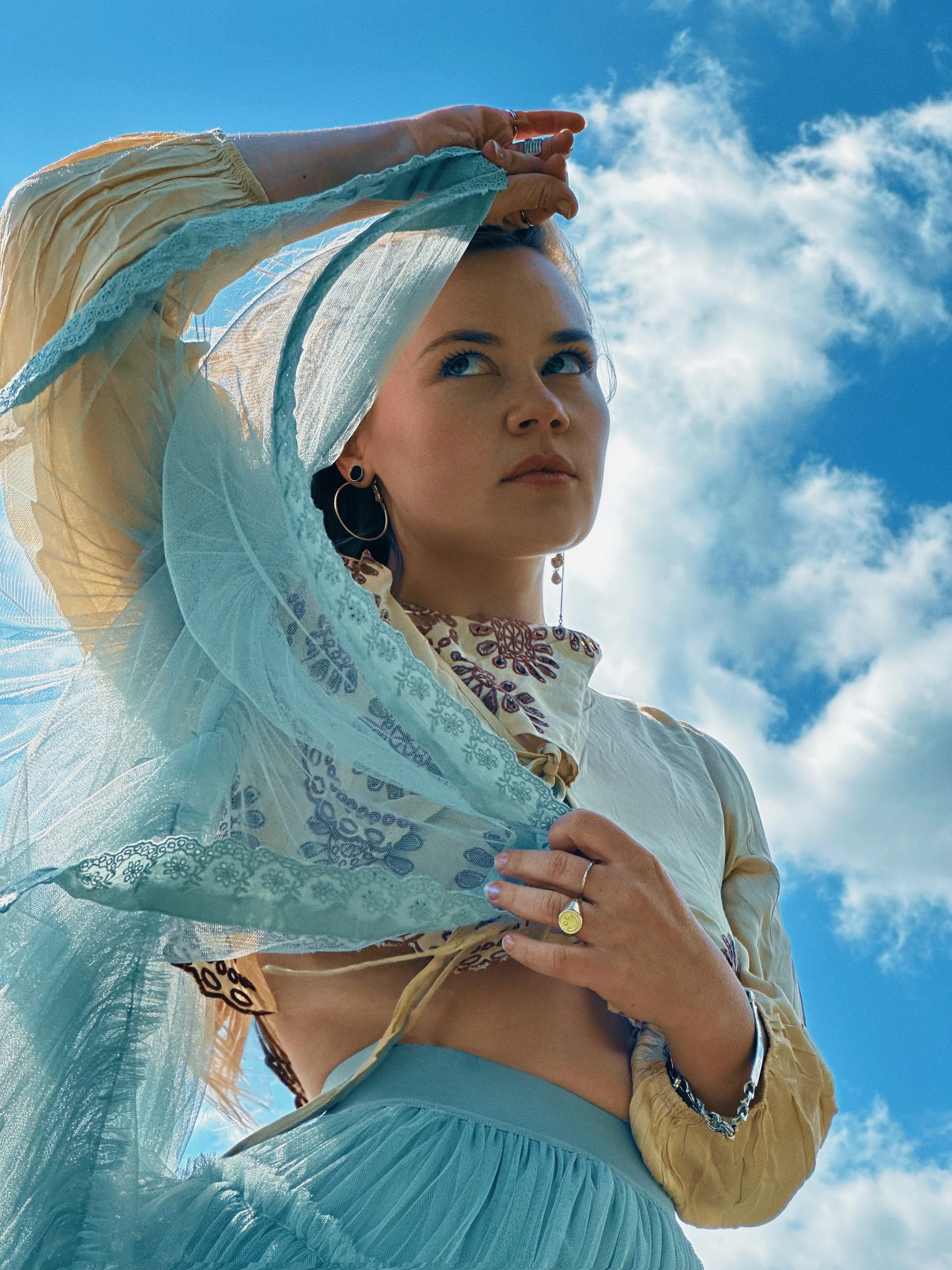 A woman looking up outdoors with a bright blue sky and scattered clouds, wearing colorful bohemian-style clothing and jewelry.