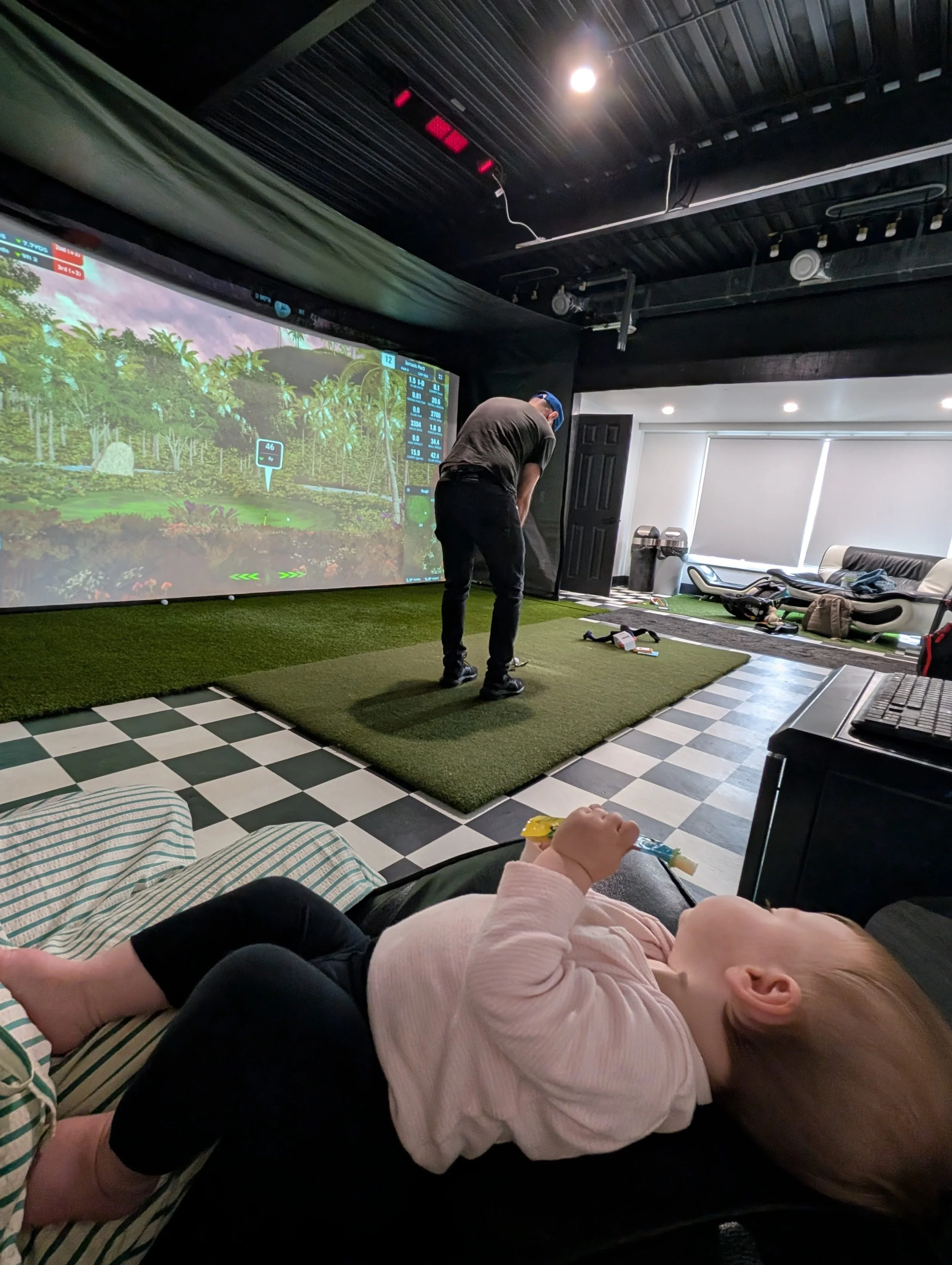 Child lying on a couch, resting or sleeping, while an adult plays golf in an indoor golf simulation room with large screen, artificial grass flooring, and gaming equipment.