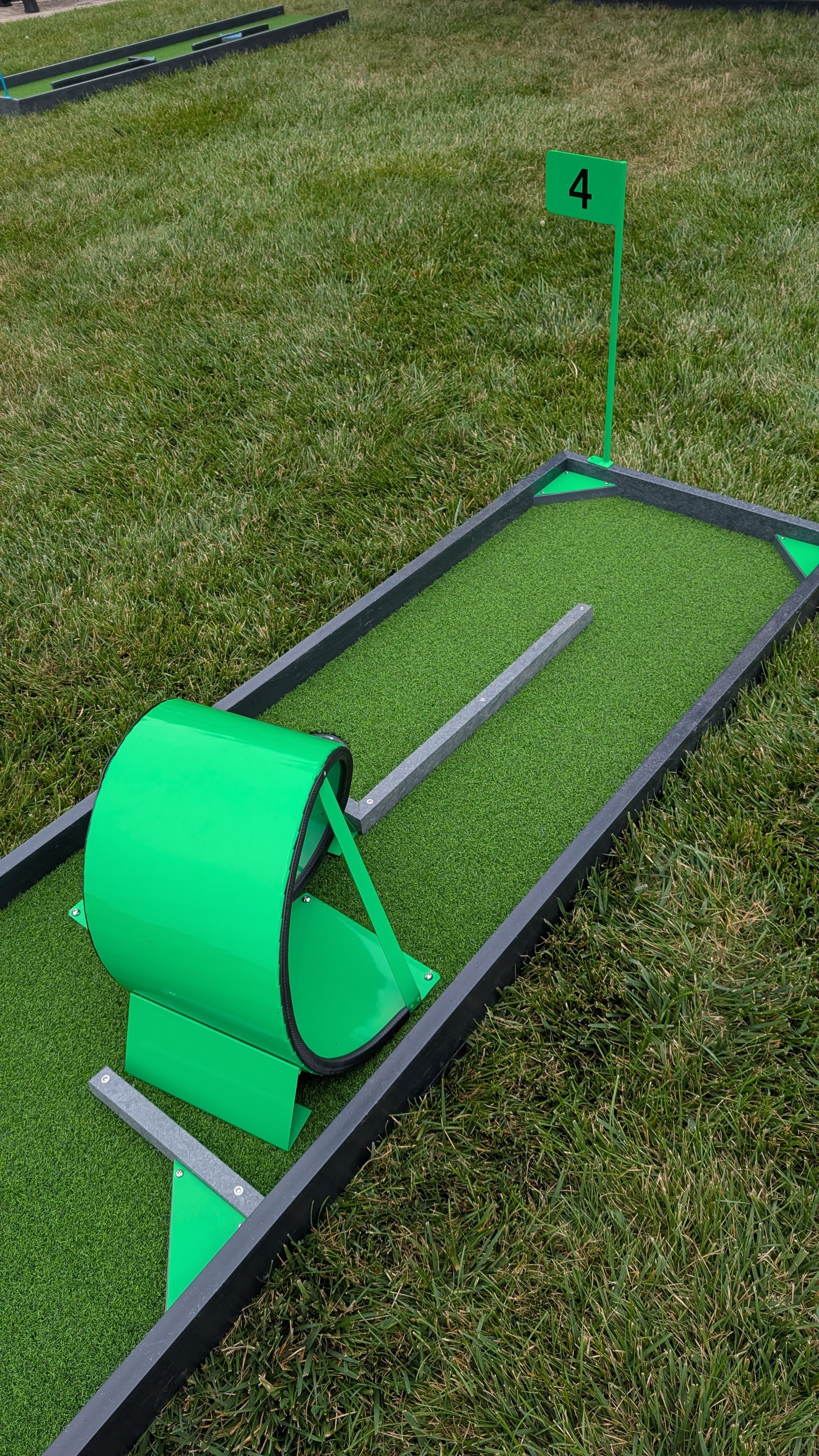A mini golf putting green with a black border, a bright green flag marked with the number 4, and a green obstacle structure on a grassy field.