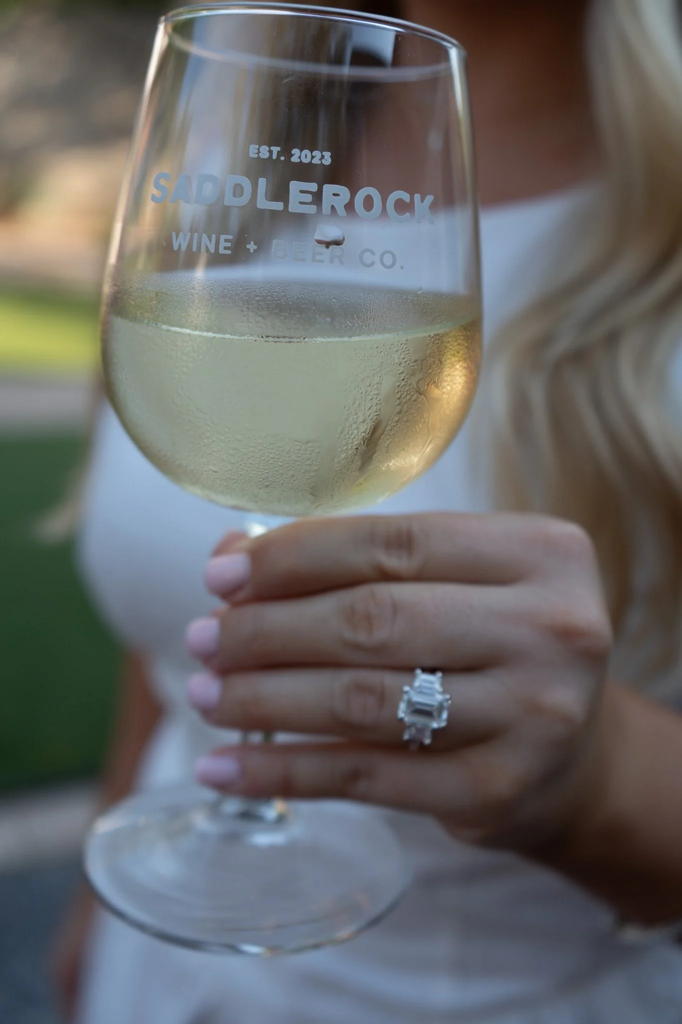 Close-up of a wine glass held by a woman, with a ring on her finger, showing the words 'SaddleRock Wine & Beer Co.' and 'Est. 2023' etched on the glass.
