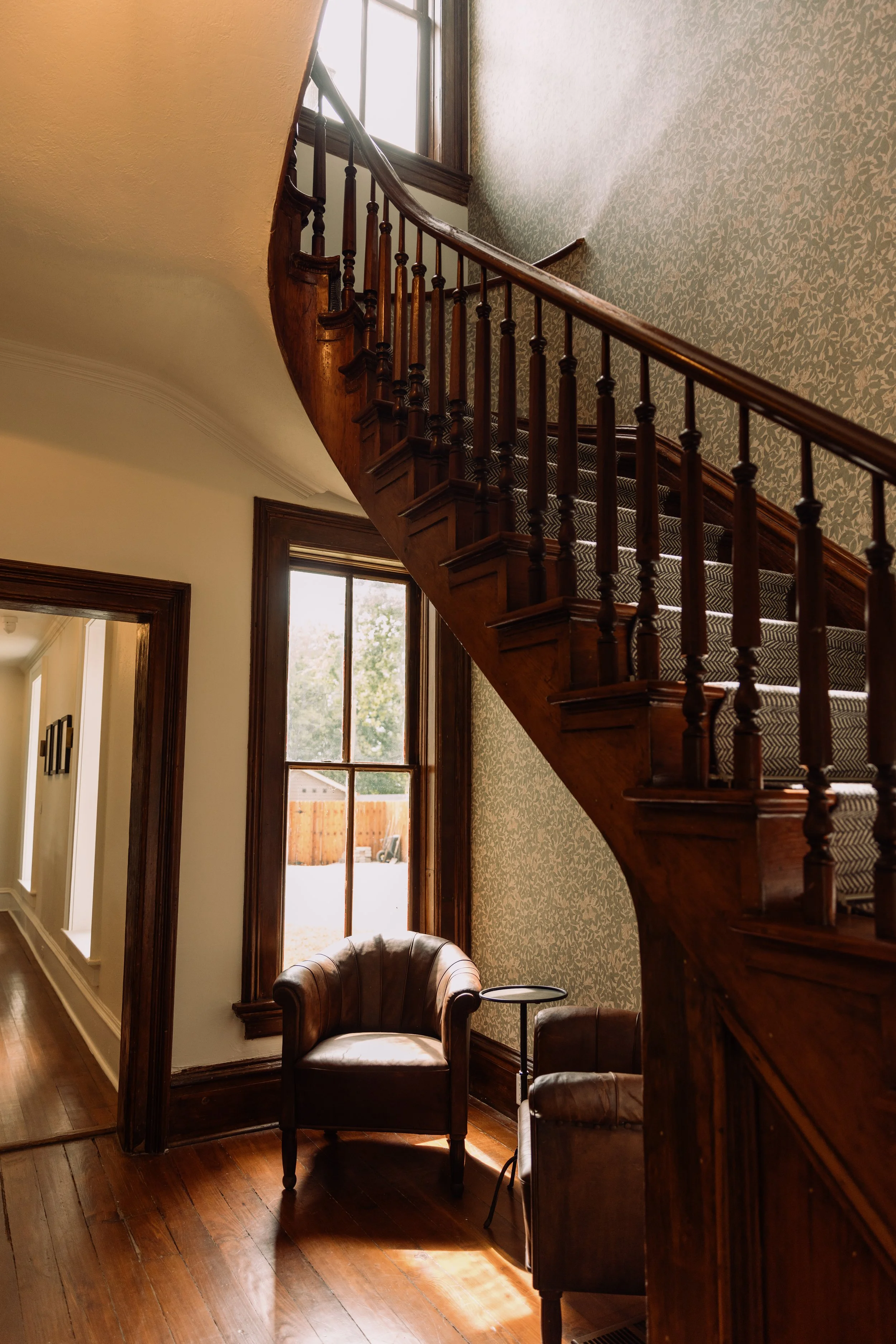 Interior view of a staircase with wooden banister, leading upstairs near large windows letting in natural light, with a sitting area of leather armchairs, wooden floors, and textured wallpaper.